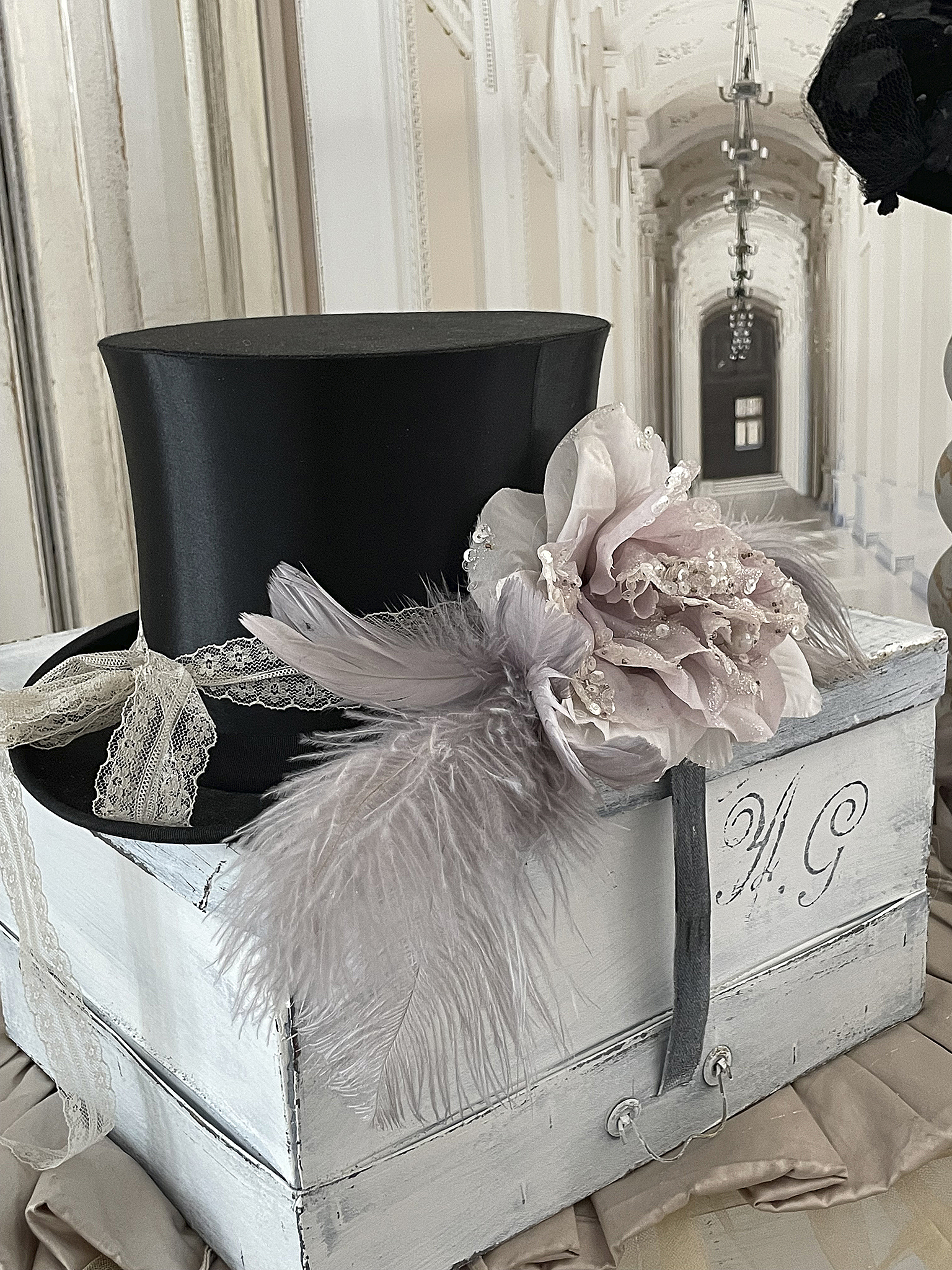 Eleganter schwarzer Zylinderhut mit zarter Blüten- und Federschmuck Schwarzer Zylinderhut dekoriert mit einem floralen Arrangement und Federn auf einem weißen Holzkoffer