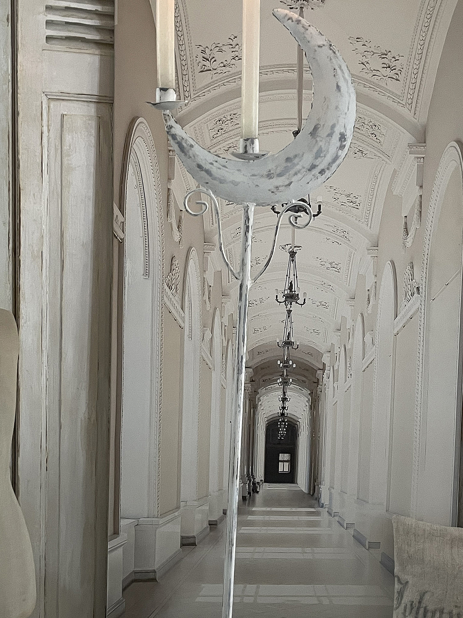 Ein eleganter Kerzenhalter in Form eines Mondes im Vordergrund einer prunkvollen, architektonisch gestalteten Halle mit langen Fluren und Kronleuchtern.