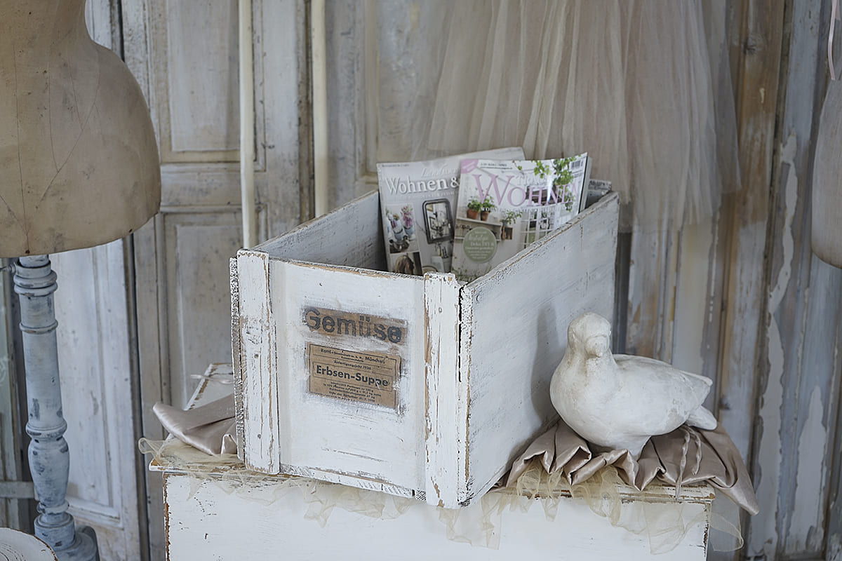 Dekorative Holzkiste mit dem Aufdruck 'Gemüse' und Zeitschriften, daneben eine weiße Deko-Taube.