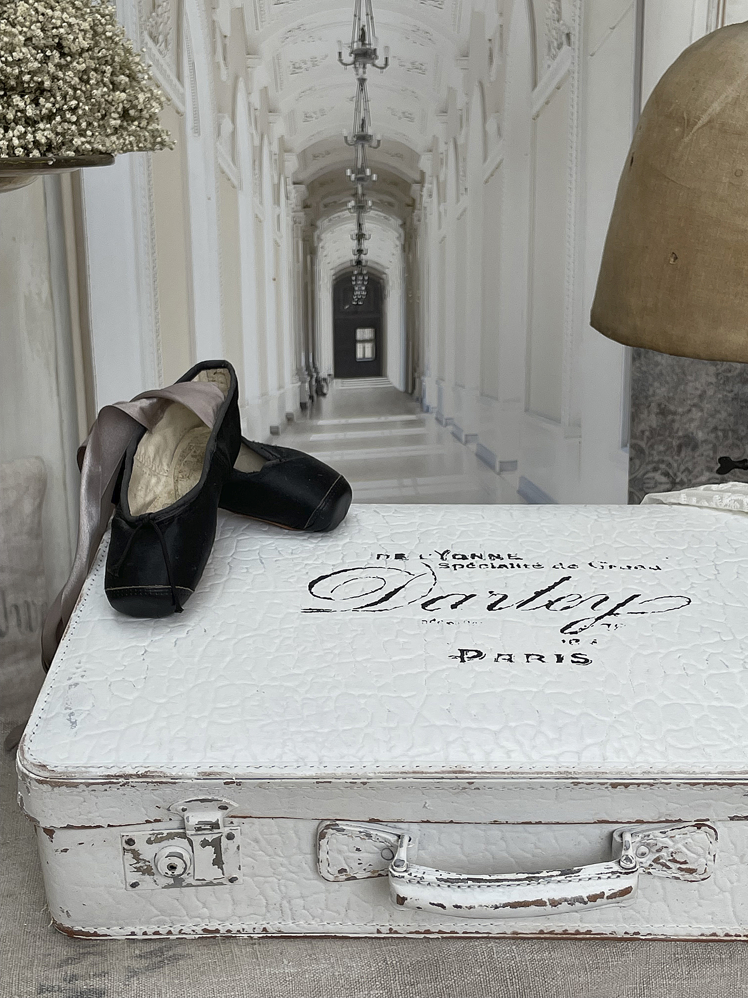 Eleganter Koffer mit Ballettschuhen und Blumendekoration Weißer Koffer mit der Aufschrift Dartey, schwarzen Ballettschuhen und einer Blumendekoration auf einem Tisch.