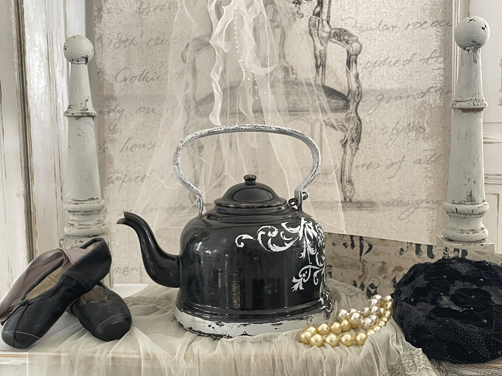 Schwarze Teekanne auf einer Tischdecke mit Ballettschuhen, Perlen und einem dekorativen Hut.
