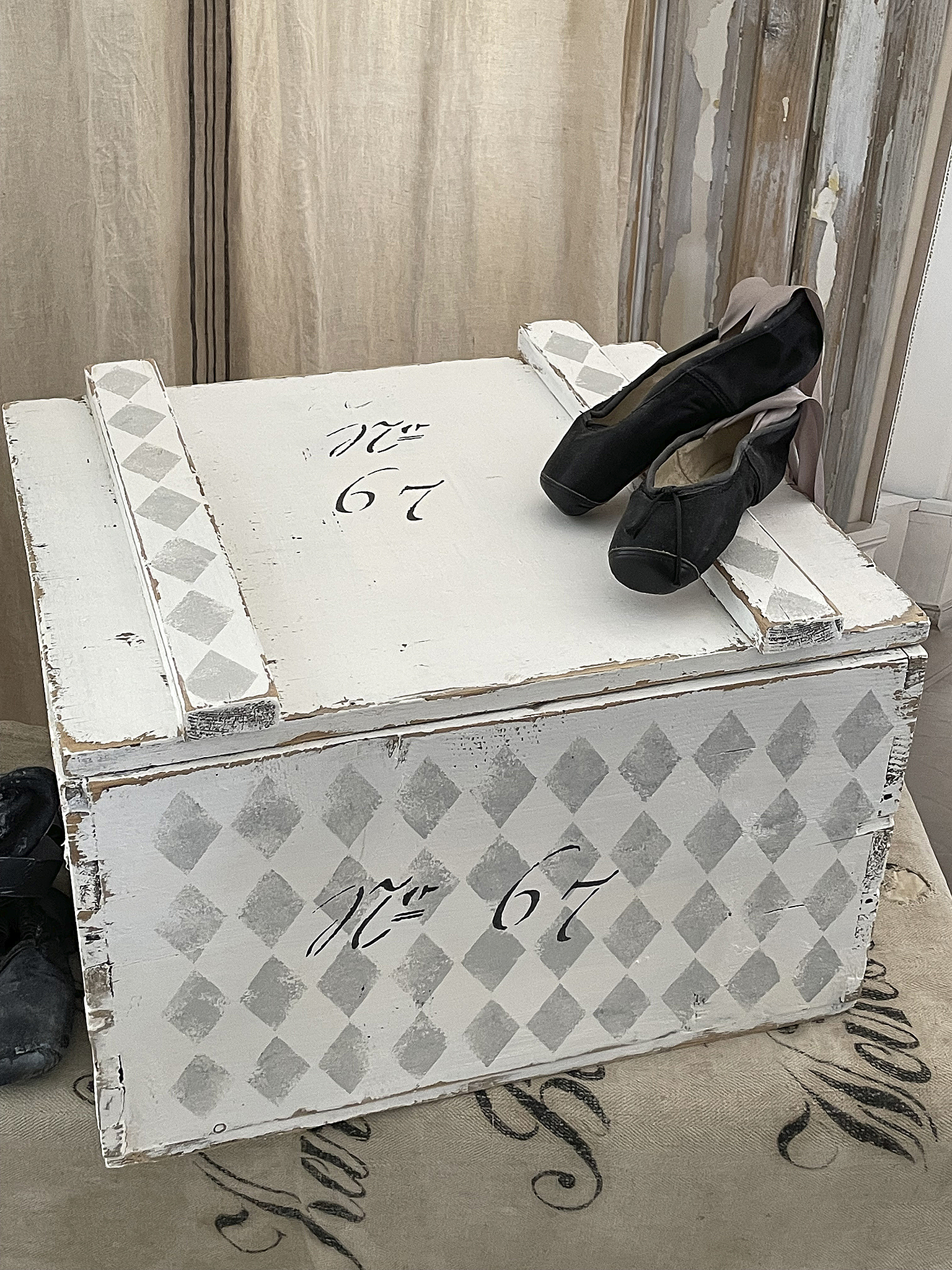Dekorative Holztruhe mit Ballettschuhen Weiße Holzkiste mit grauen Rauten und der Nummer 67, darauf liegen schwarze Ballettschuhe.