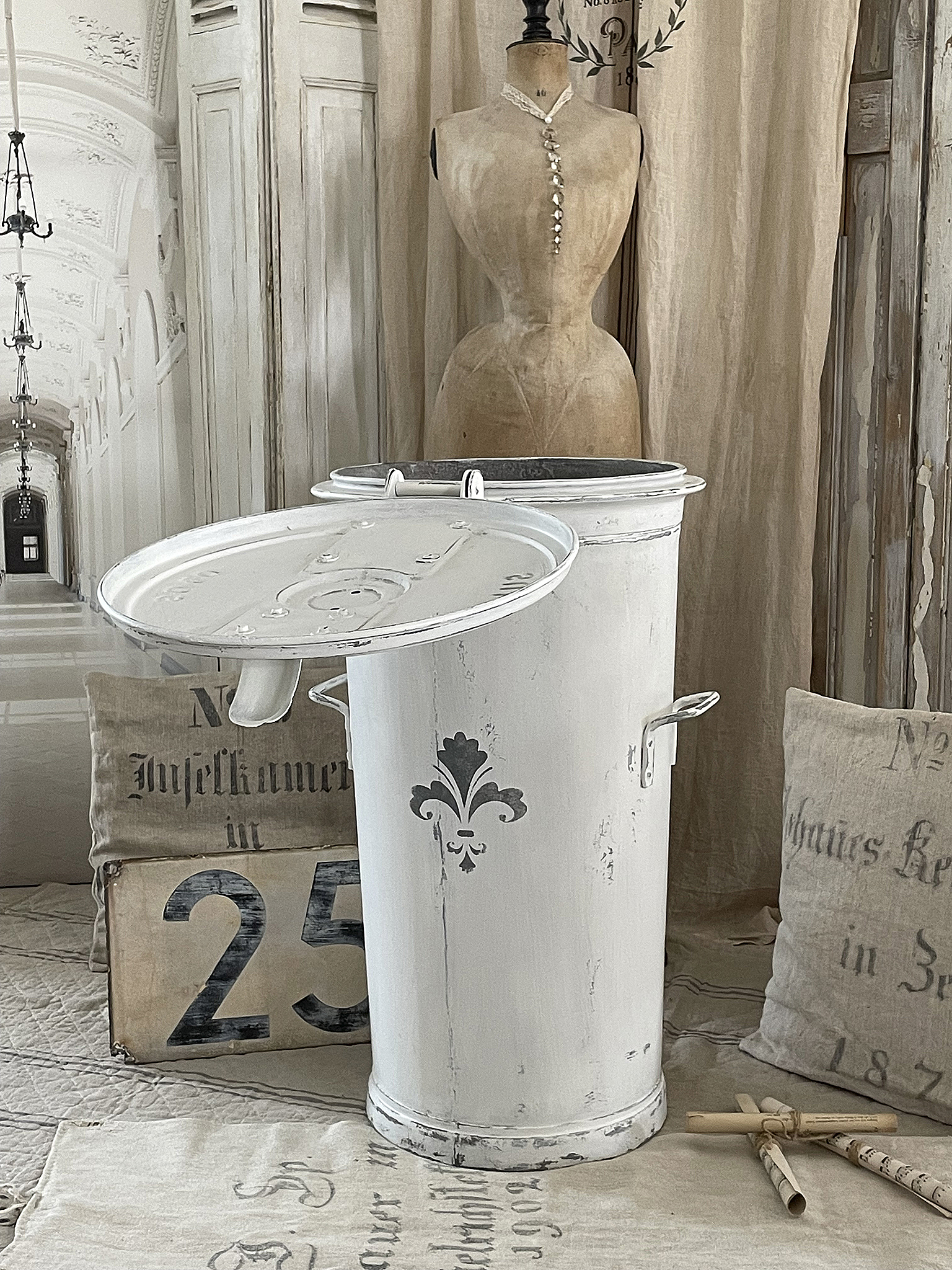 Klassischer Vintage-Mülleimer in Weiß mit Fleur-de-Lis-Muster Weißer Vintage-Mülleimer mit aufklappbarem Deckel und elegantem Design, im Hintergrund eine Schneiderpuppe.