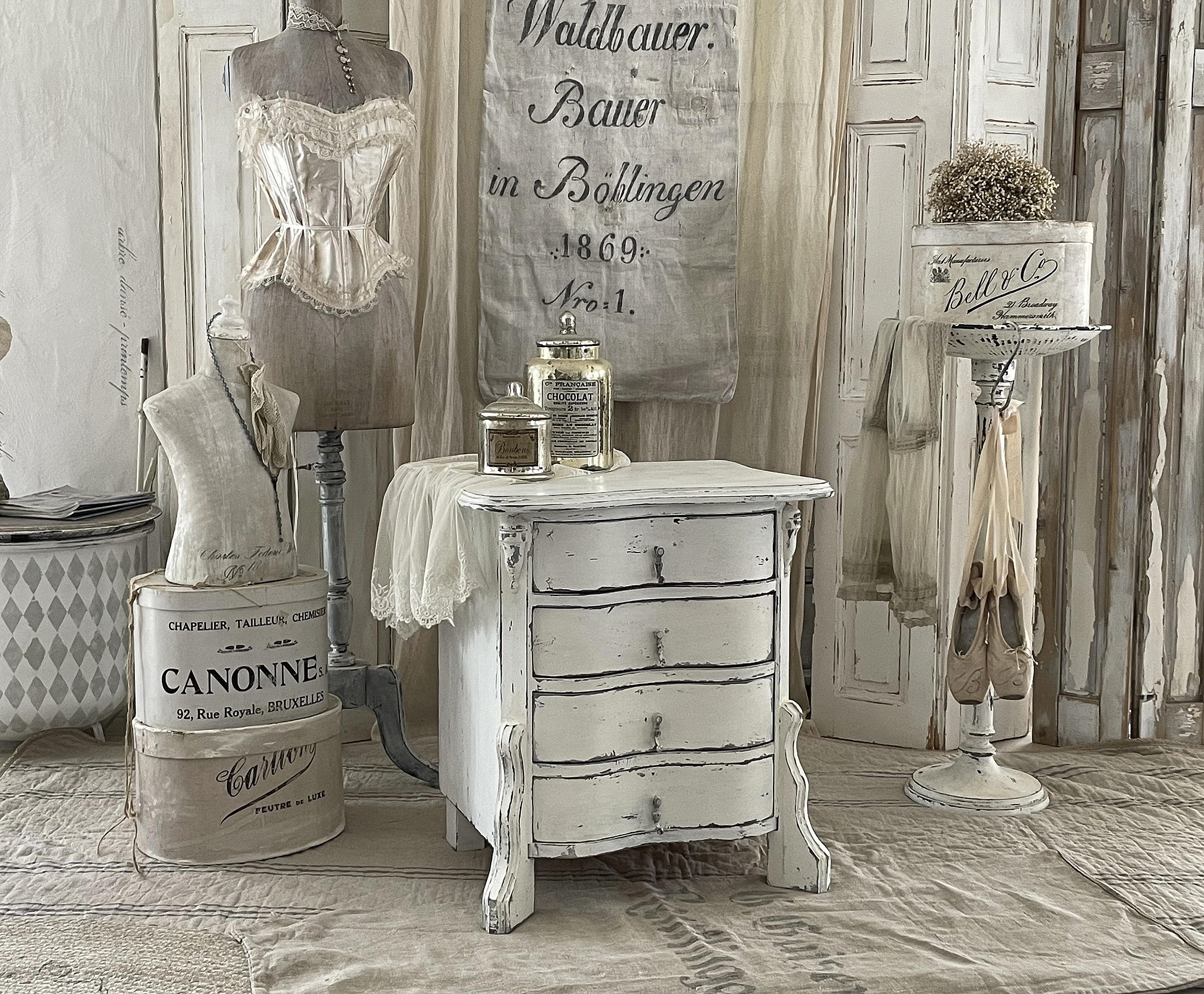 Klassischer Vintage-Tisch mit viel Charme und romantischen Accessoires Ein nostalgischer Raum mit einem weißen Vintage-Nachttisch, einer Schneiderpuppe und eleganten Deko-Elementen.