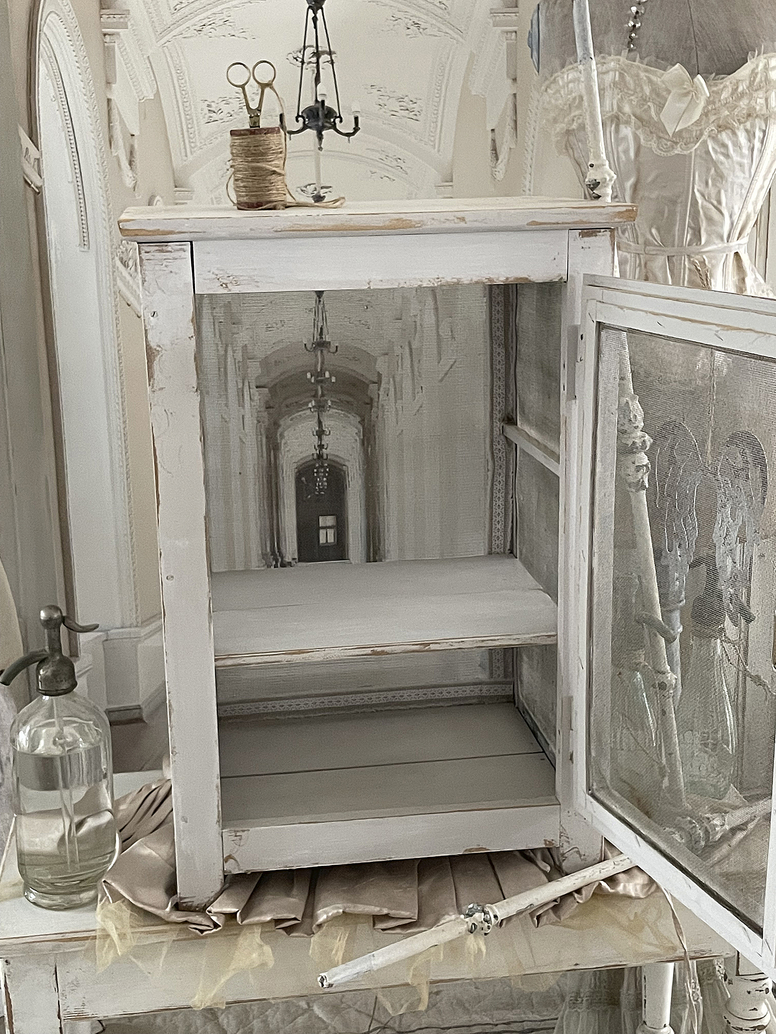 Schöner Vintage-Kasten mit offenen Türen und dekorativen Elementen Vintage-Kasten in Weiß mit zwei Regalen und einer offenen Tür, dekoriert mit Scheren und Garn in einem eleganten Raum.