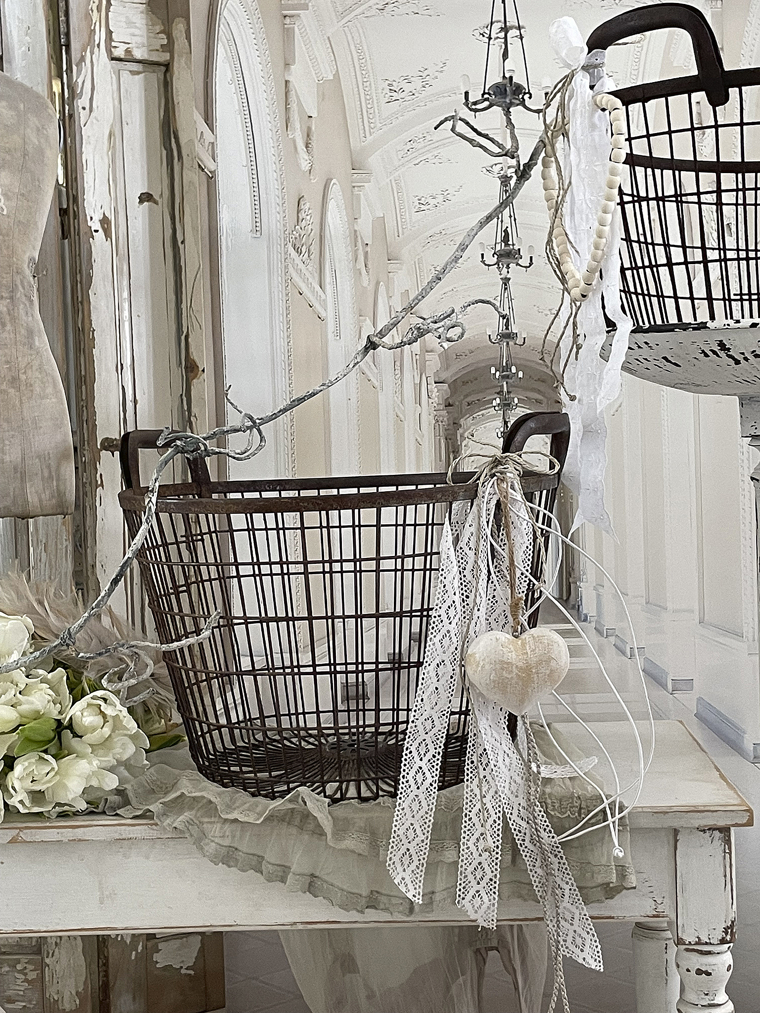 Dekorativer Drahtkorb mit Herz und Bändern auf einem Tisch Rustikaler Drahtkorb mit herzförmigem Anhänger und floralen Elementen, dekoriert im Shabby-Chic-Stil.