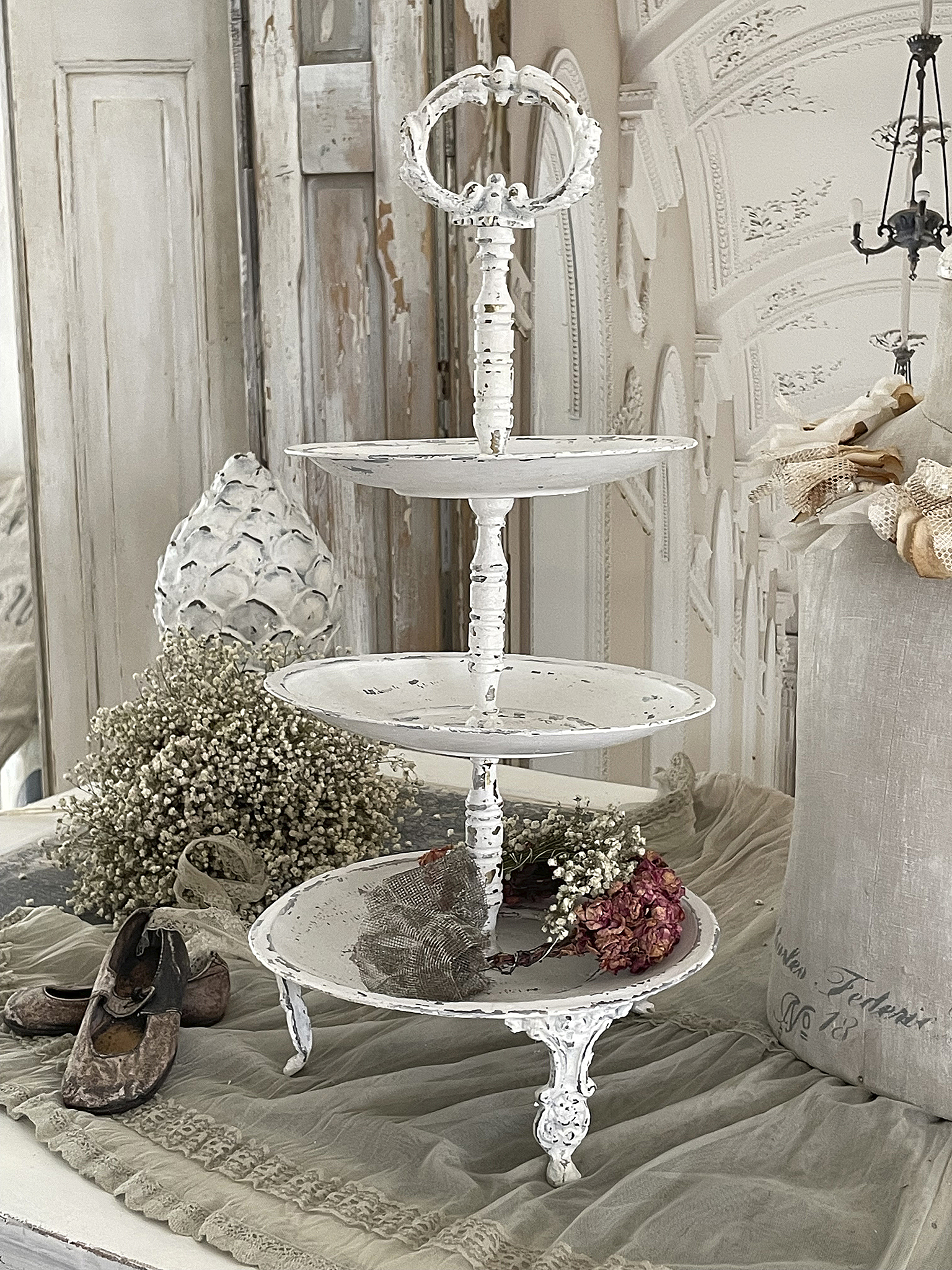 Eleganter weißer Etagere mit Blumenarrangement Weißer Etagere mit drei Etagen, umgeben von Trockenblumen und alten Schuhen auf einem beige Tisch
