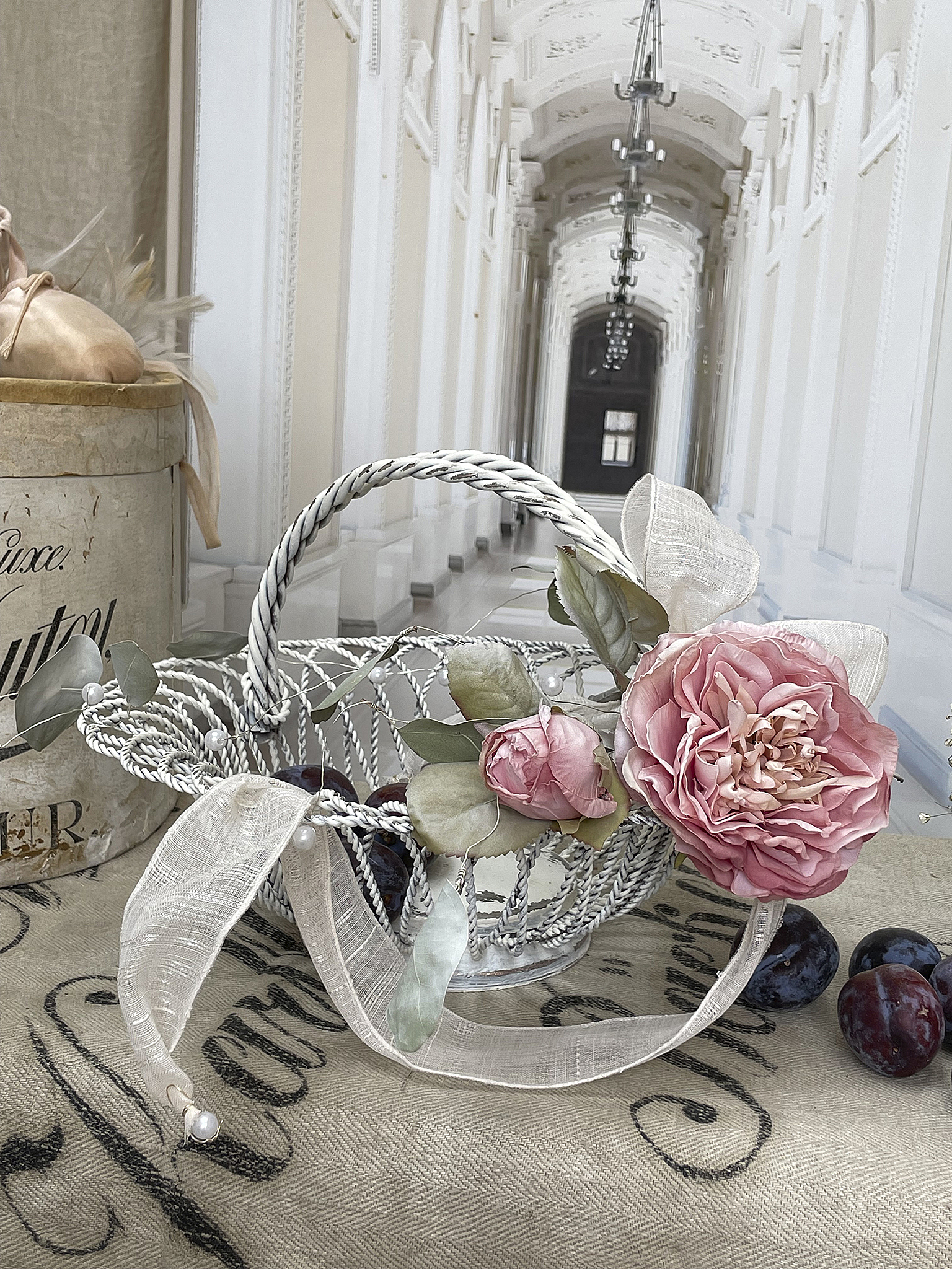 Eleganter Korb mit künstlichen Rosen und frischen Pflaumen, ideal für Dekoration. Dekorativer Korb mit Rosen und Pflaumen auf einem vintage Tisch. Im Hintergrund ein eleganter Gang.