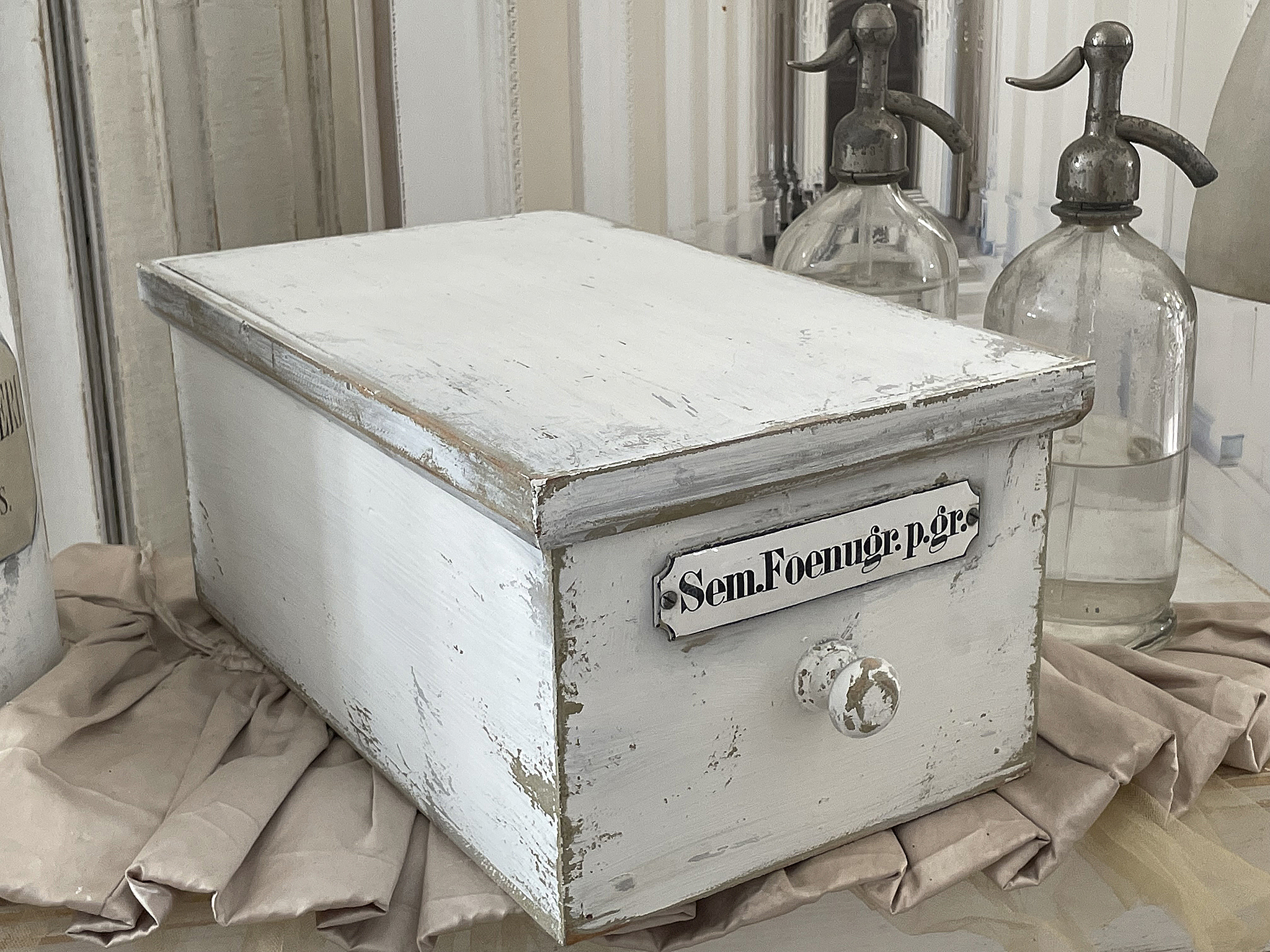 Historische Aufbewahrungsbox in Weiß mit Beschriftung Vintage Holzbox mit der Aufschrift 'Sem.Foenugr.p.gr.' auf einer dekorativen Tischunterlage.