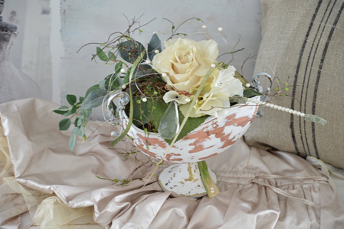 Blumenarrangement mit rosafarbener Rose und Dekoelementen in antikem Gefäß auf cremefarbenem Stoff