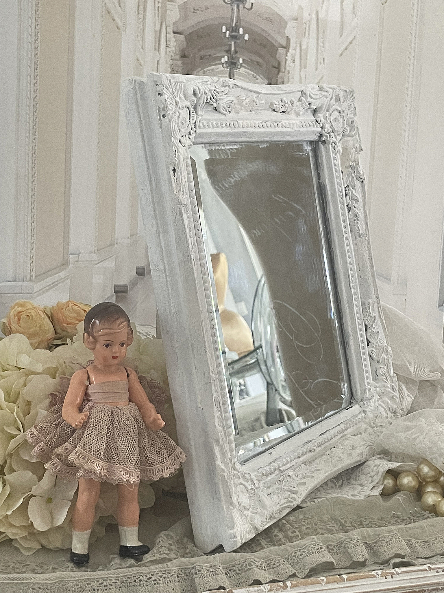 Vintage Puppe und Spiegel in stilvollem Arrangement Eine antike Puppe in einem zarten Kleid steht neben einem verzierten Spiegel auf einem dekorativen Tisch.