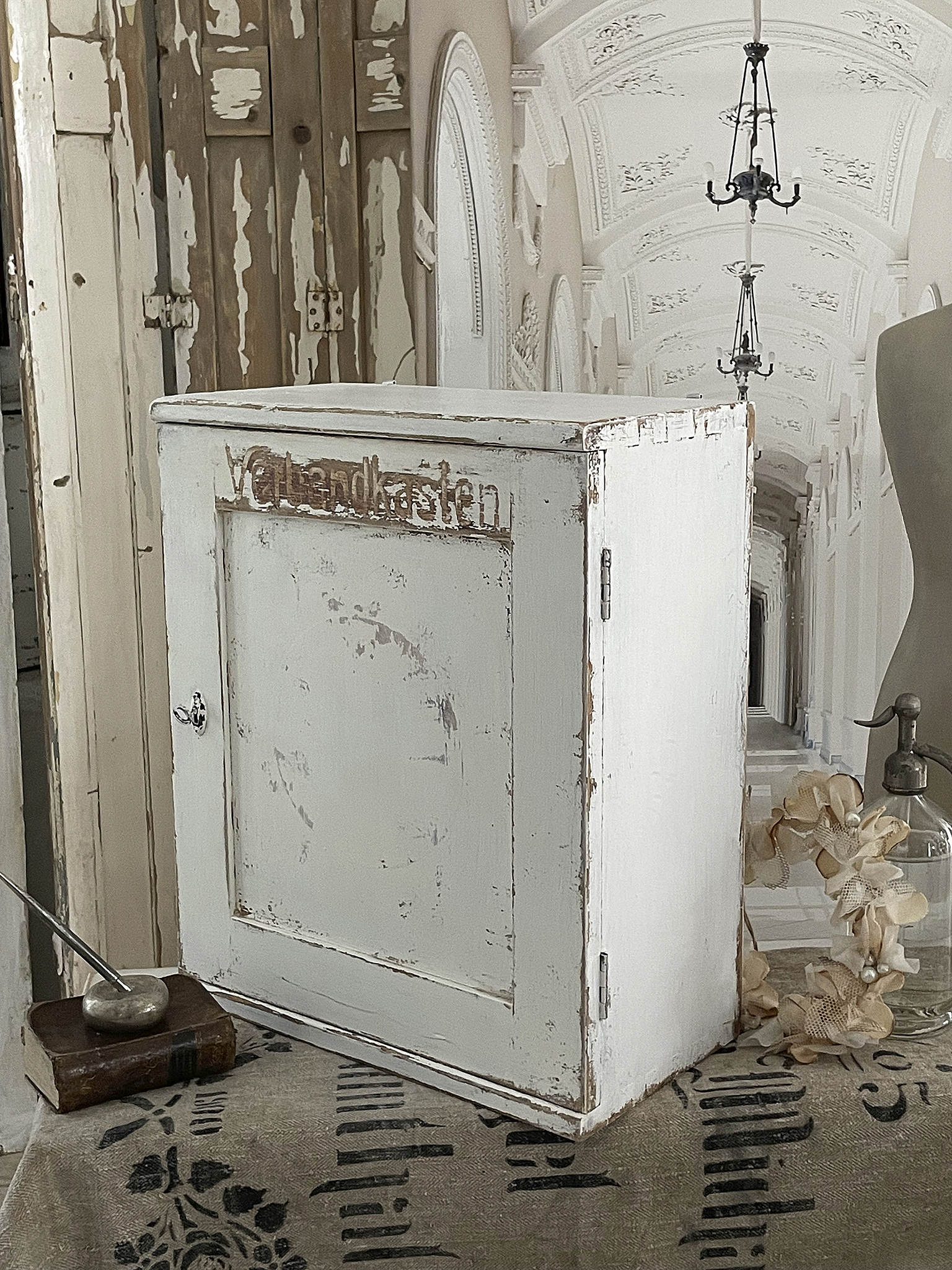 Vintage weißer Schrank mit Beschriftung "Verbandkasten" im nostalgischen Ambiente Nostalgischer Schrank in Weiß mit der Aufschrift "Verbandkasten" vor einer dekorativen Kulisse