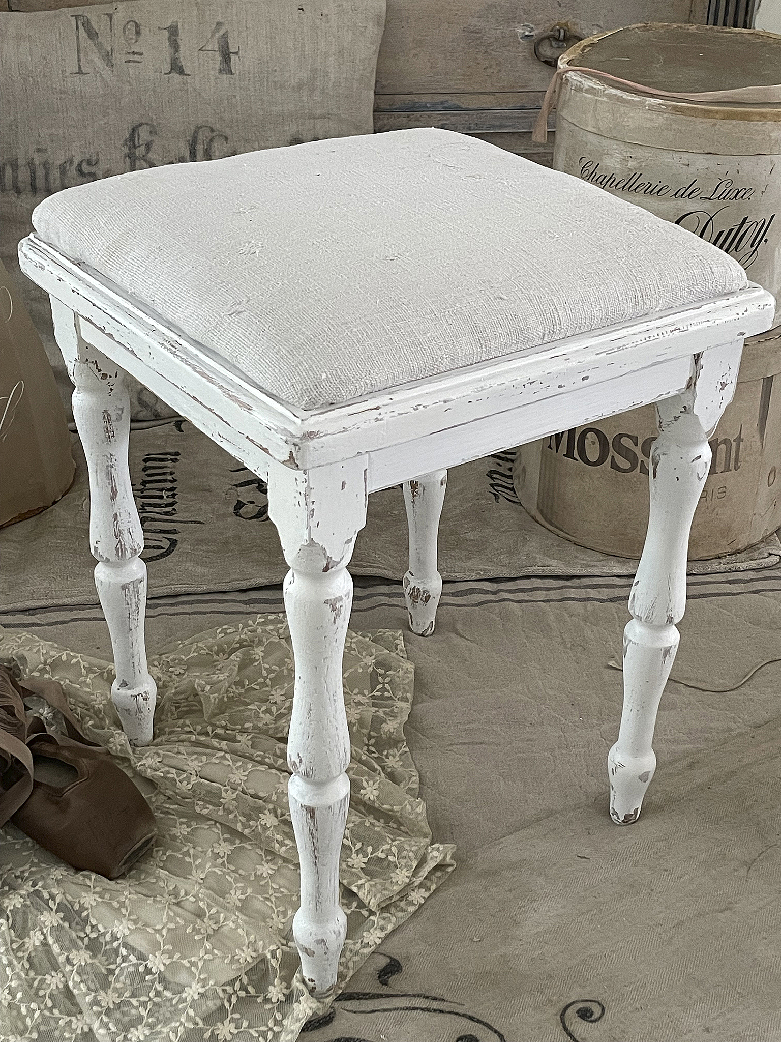 Eleganter weißer Hocker mit gepolsterter Sitzfläche Weißer Hocker mit abgenutztem Look und gepolsterter Sitzfläche, dekorativ platziert.
