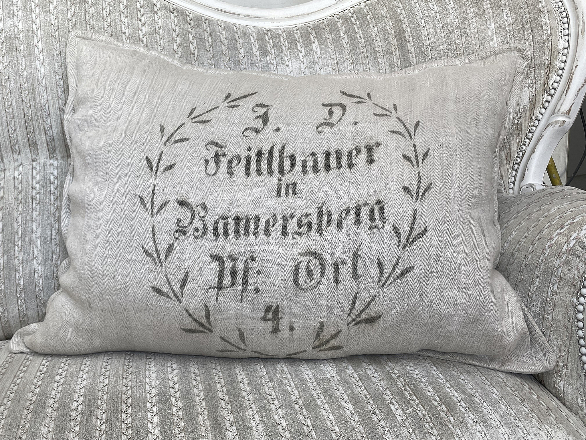 Dekoratives Kissen mit nostalgischem Aufdruck aus Bannersberg Kissen mit dem Aufdruck 'J. D. Feiltbauer in Bannersberg Pf. Ort 4' auf einem gepolsterten Stuhl.