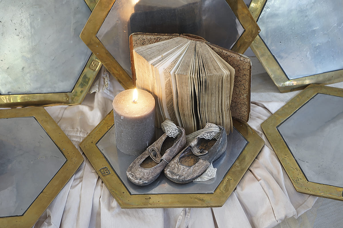 Detailaufnahme von antiken Babyschuhen, einer brennenden Kerze und einem alten Buch auf hexagonalen Spiegeln.