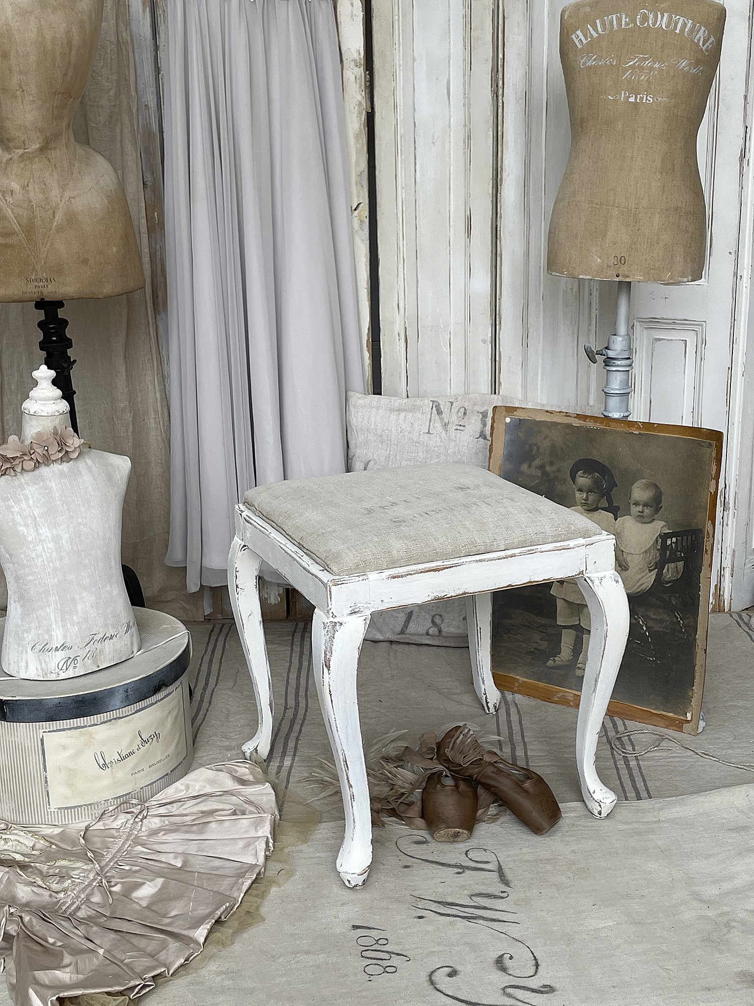 Schöner Vintage Hocker mit abgenutzter weißer Farbe, umgeben von Mannequins und nostalgischen Accessoires.
