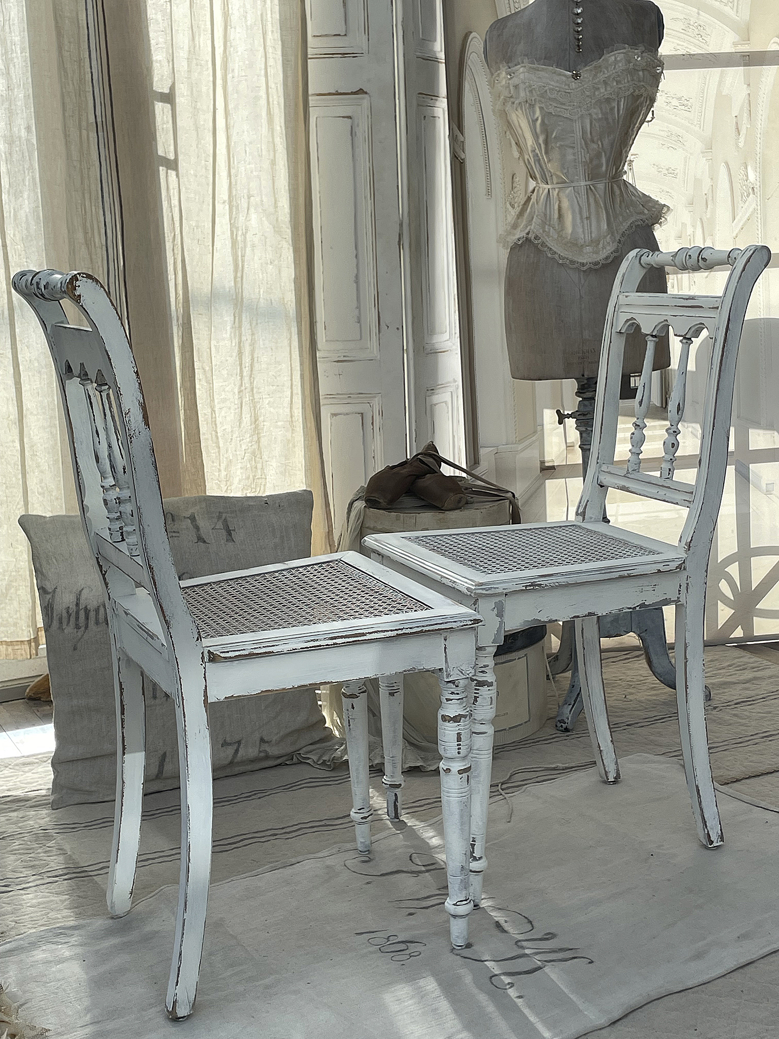Zwei weiße Shabby-Chic-Stühle in einem hellen Raum, umgeben von Stoffen und einer Schneiderpuppe.