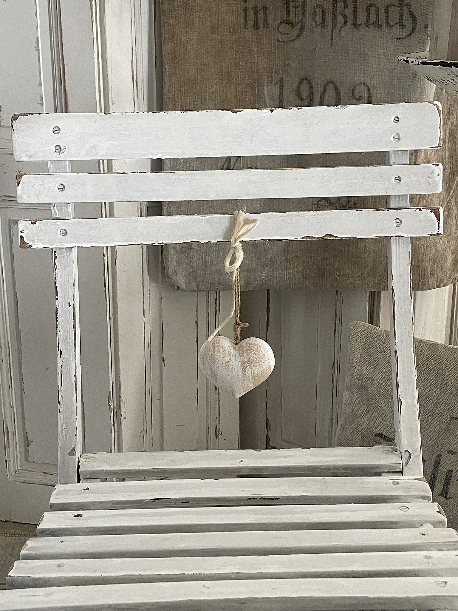 Weißer Holzstuhl mit herzförmigem Dekoelement Ein veralteter weißer Holzstuhl mit einem hängenden herzförmigen Ornament.