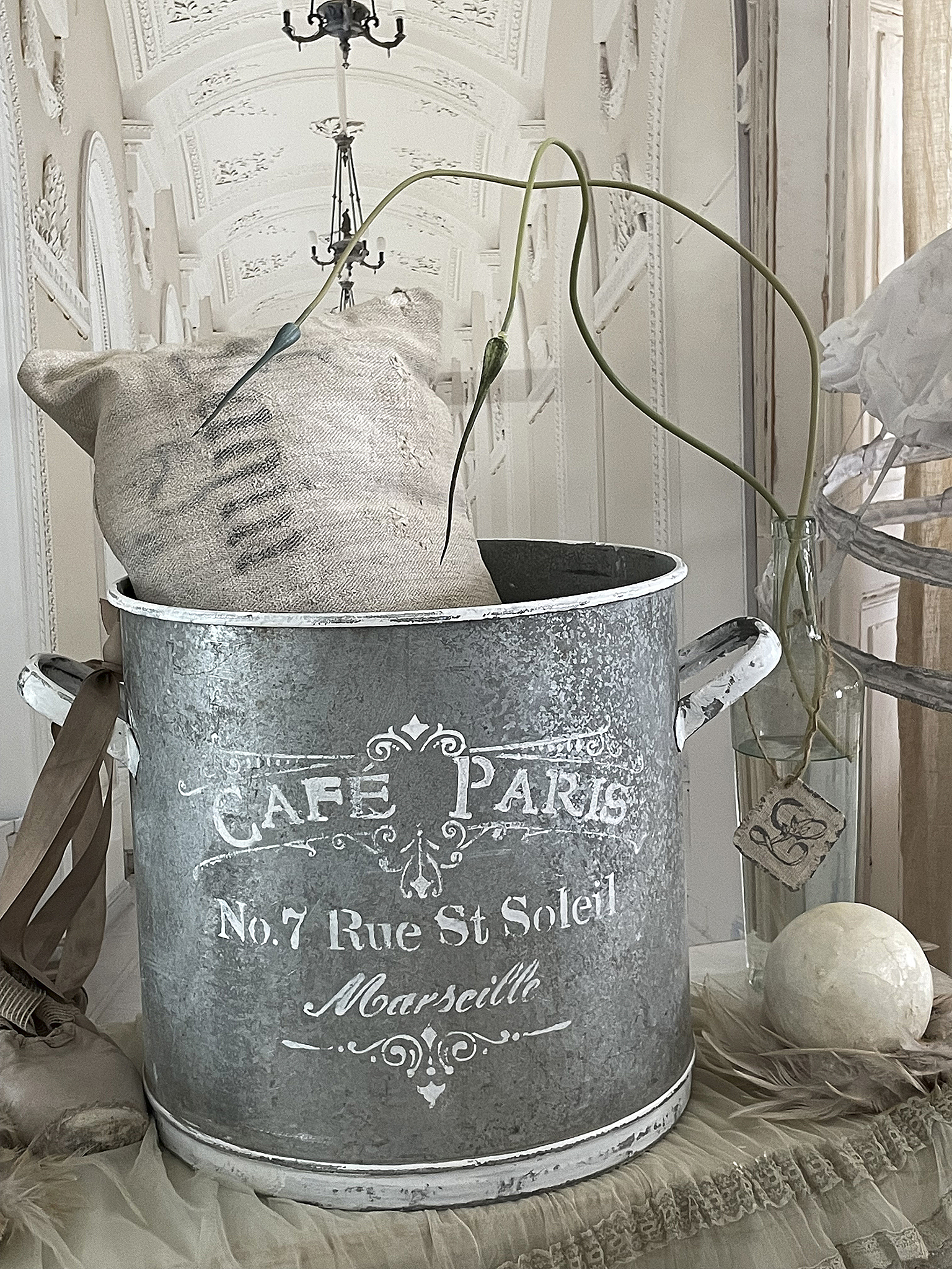 Dekorativer Metallbehälter mit Aufschrift und Pflanzen Älterer Metallbehälter mit Aufschrift 'Cafe Paris No.7 Rue St Soleill Marseille', gefüllt mit einem Sack und einer Pflanze.