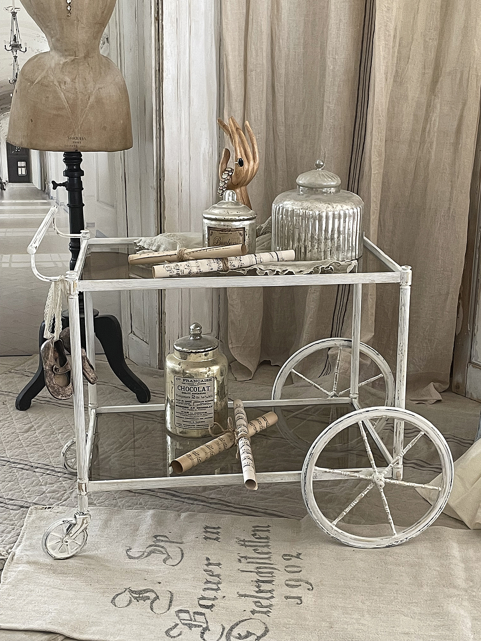 Eleganter Servierwagen mit Vintage-Dekoration und stilvollen Accessoires Servierwagen in Weiß mit Glasbehältern, Notenrollen und einer Schneiderpuppe im Hintergrund