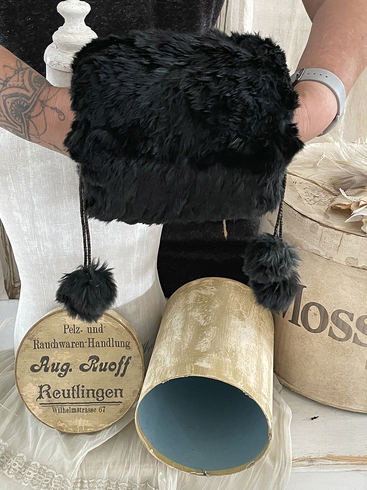 Schwarze Pelzmütze mit hängenden Pompons und vintage Hutbox Schwarze Pelzmütze, die von einer Person gehalten wird, zusammen mit einer vintage Hutbox von Aug. Ruoff, Reutlingen.