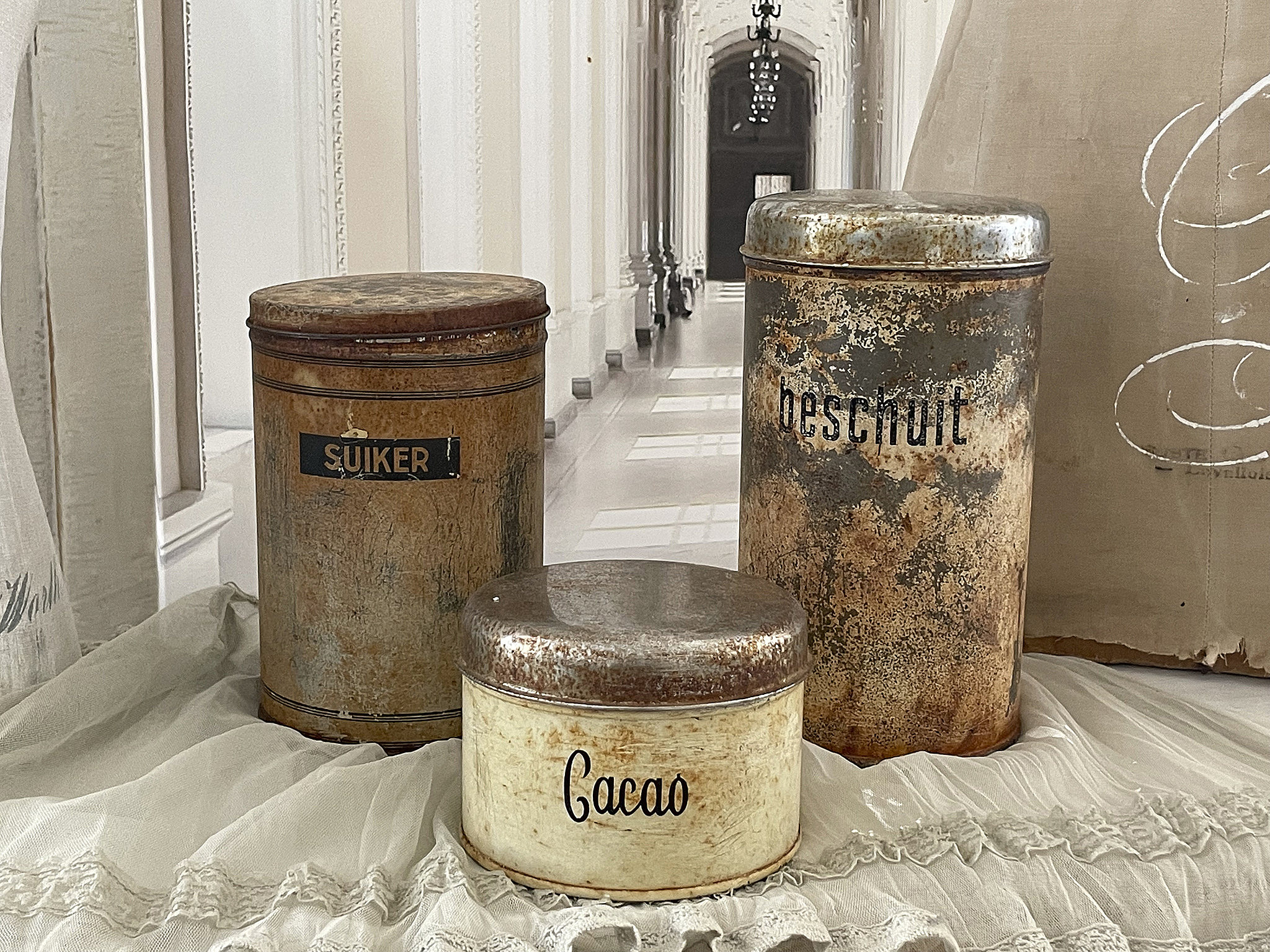 Zwei Vintage-Dosen aus Metall mit Beschriftungen Drei antike Metalldosen mit Beschriftungen wie Suiker, Beschuit und Cacao auf einem gedeckten Tisch