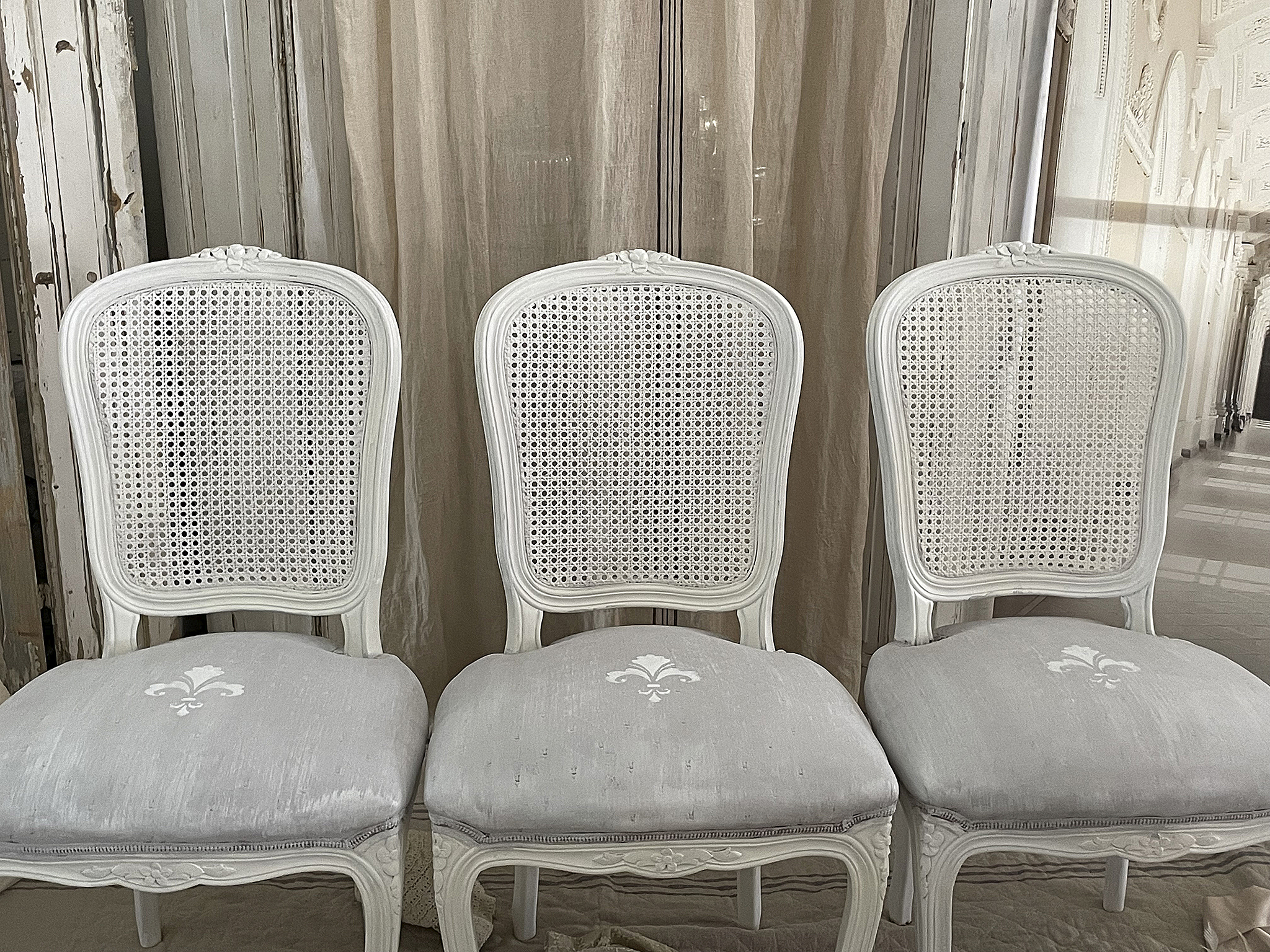 Elegante weiße Stühle mit Rattangeflecht und grauen Polstern Drei stilvolle weiße Stühle mit Rattangeflecht und grauen Stoffbezügen im Hintergrund