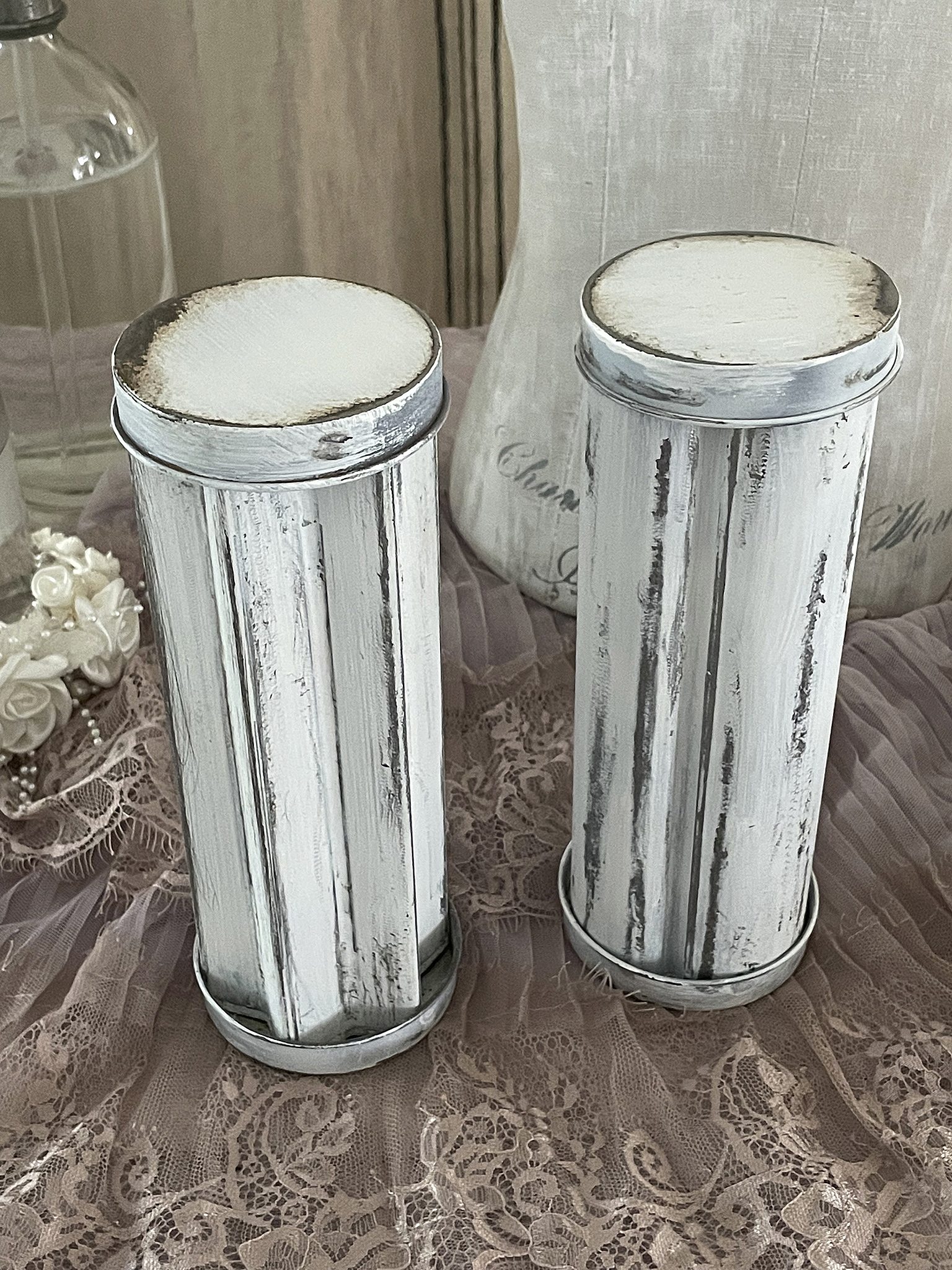 Zwei vintage inspirierte Metall-Dosen mit weißer Farbe und antikem Finish auf einem Tisch mit Spitze.