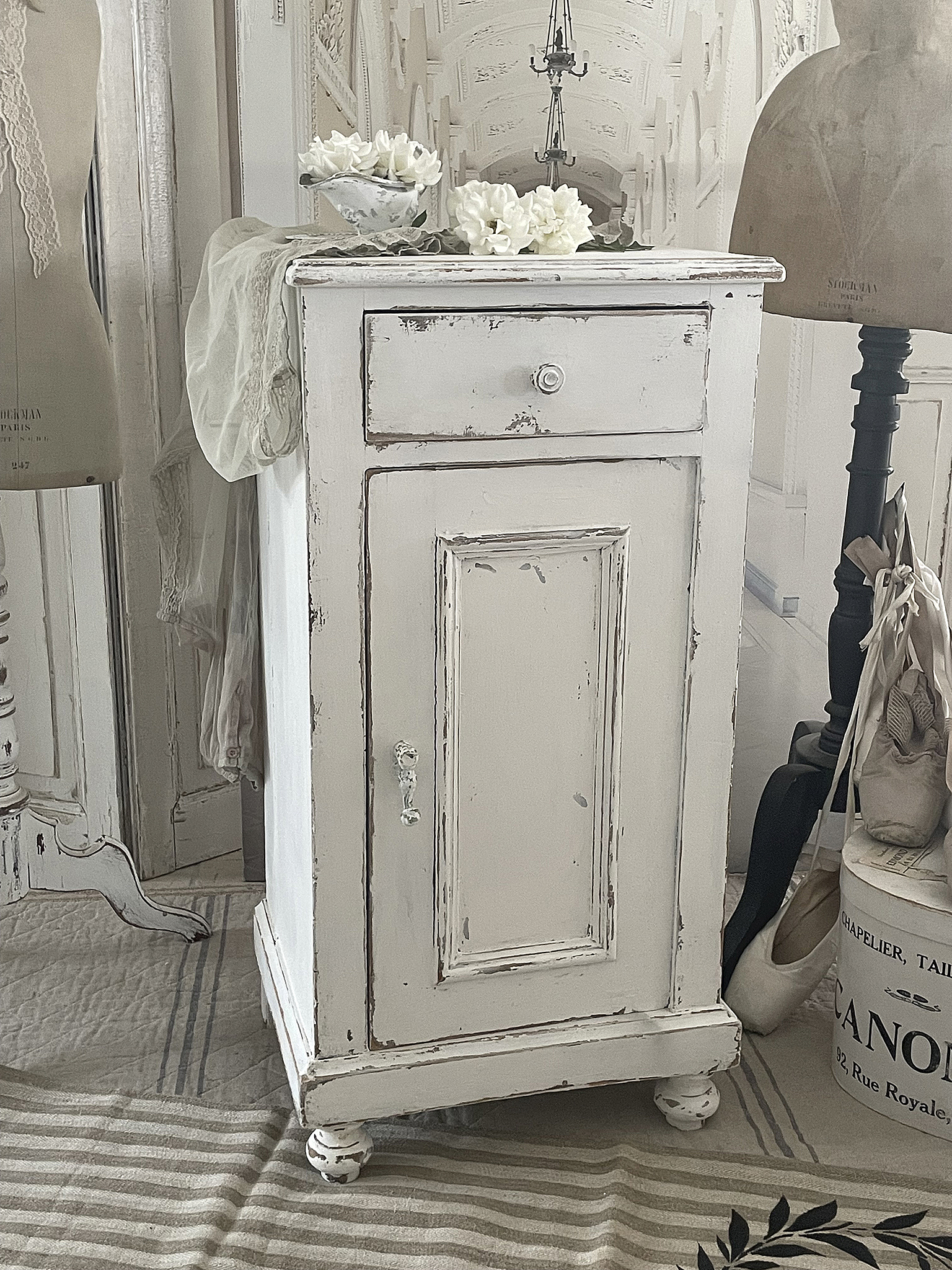 Eleganter Shabby Chic Nachtschrank mit einer Schublade Shabby Chic Nachtschrank in Weiß mit Blüten und Spiegel im Hintergrund