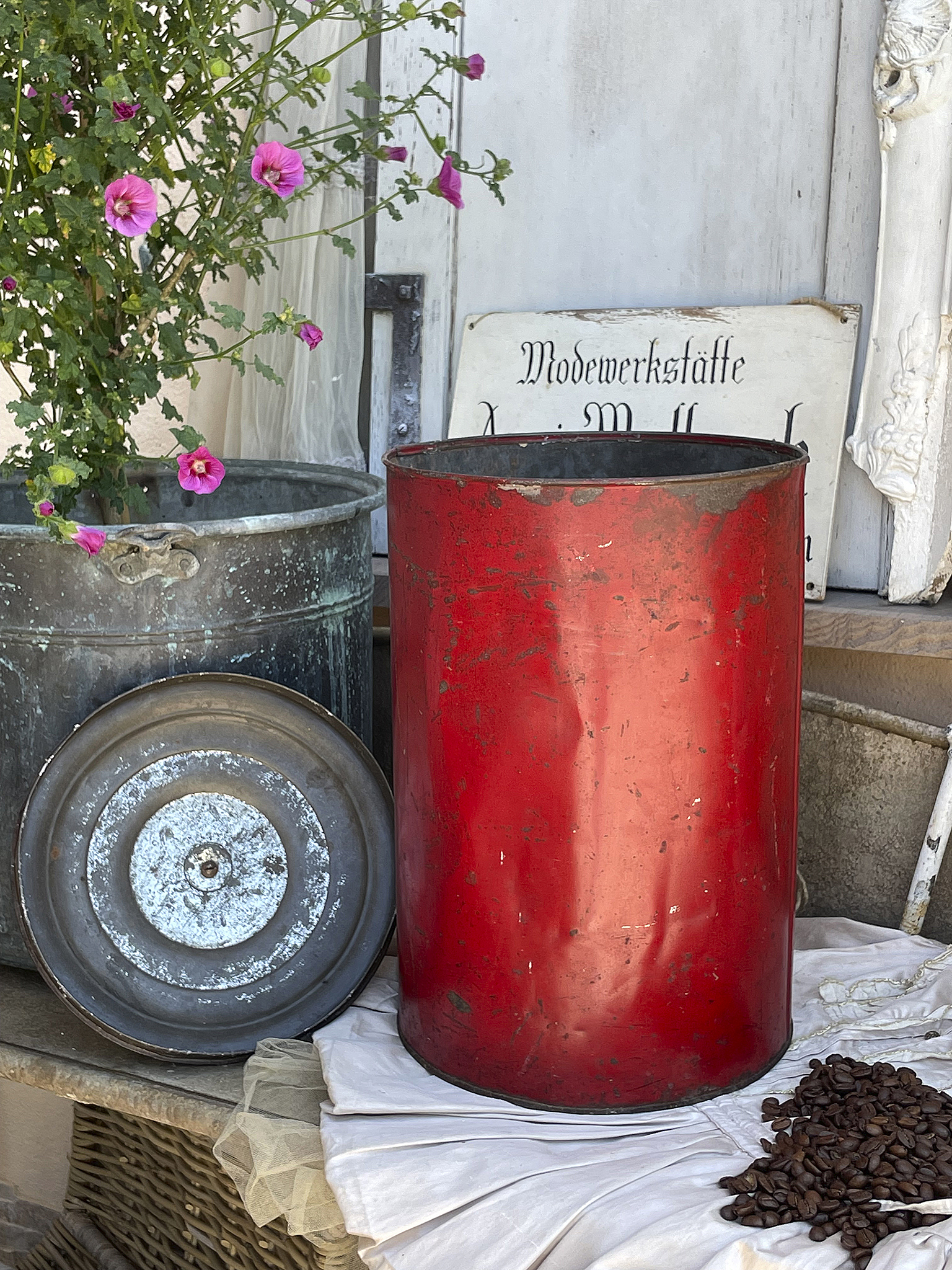 Dekorativer roter Kanister mit Blumentopf und Kaffeebohnen Roter Kanister, daneben ein grauer Behälter, umgeben von Blüten und Kaffeebohnen.