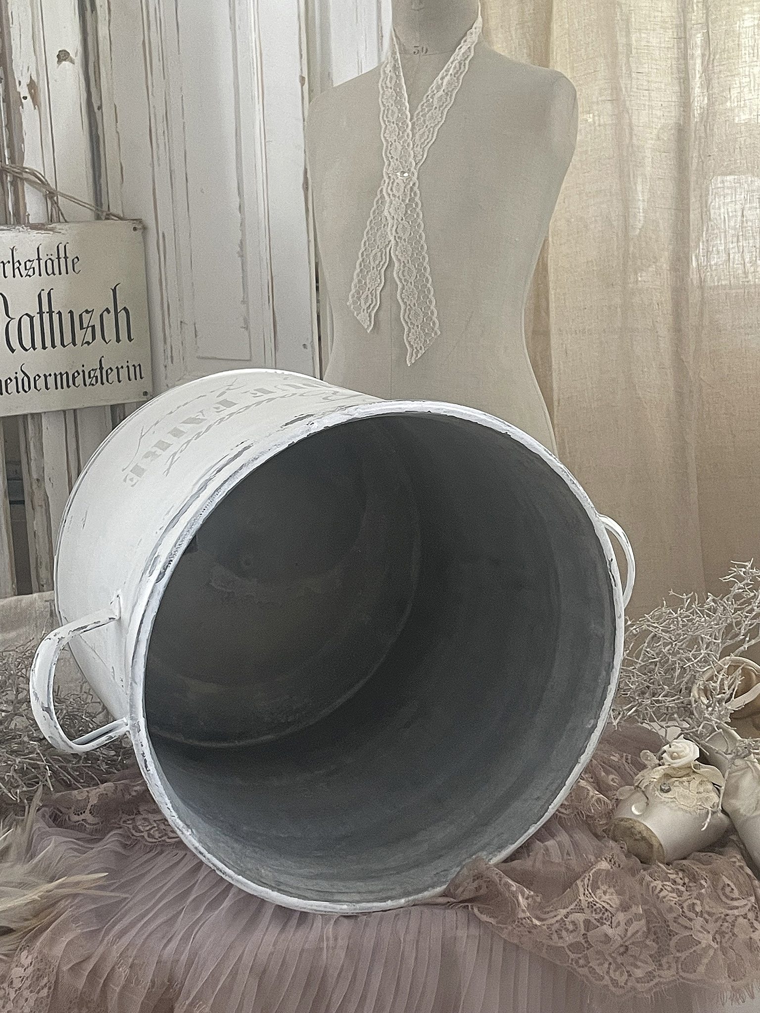 Hübscher weißer Metall-Eimer mit Vintage-Charme Vielseitiger weißer Metall-Eimer auf einem rosa Laken, dekoriert mit Spitze und romantischen Accessoires.