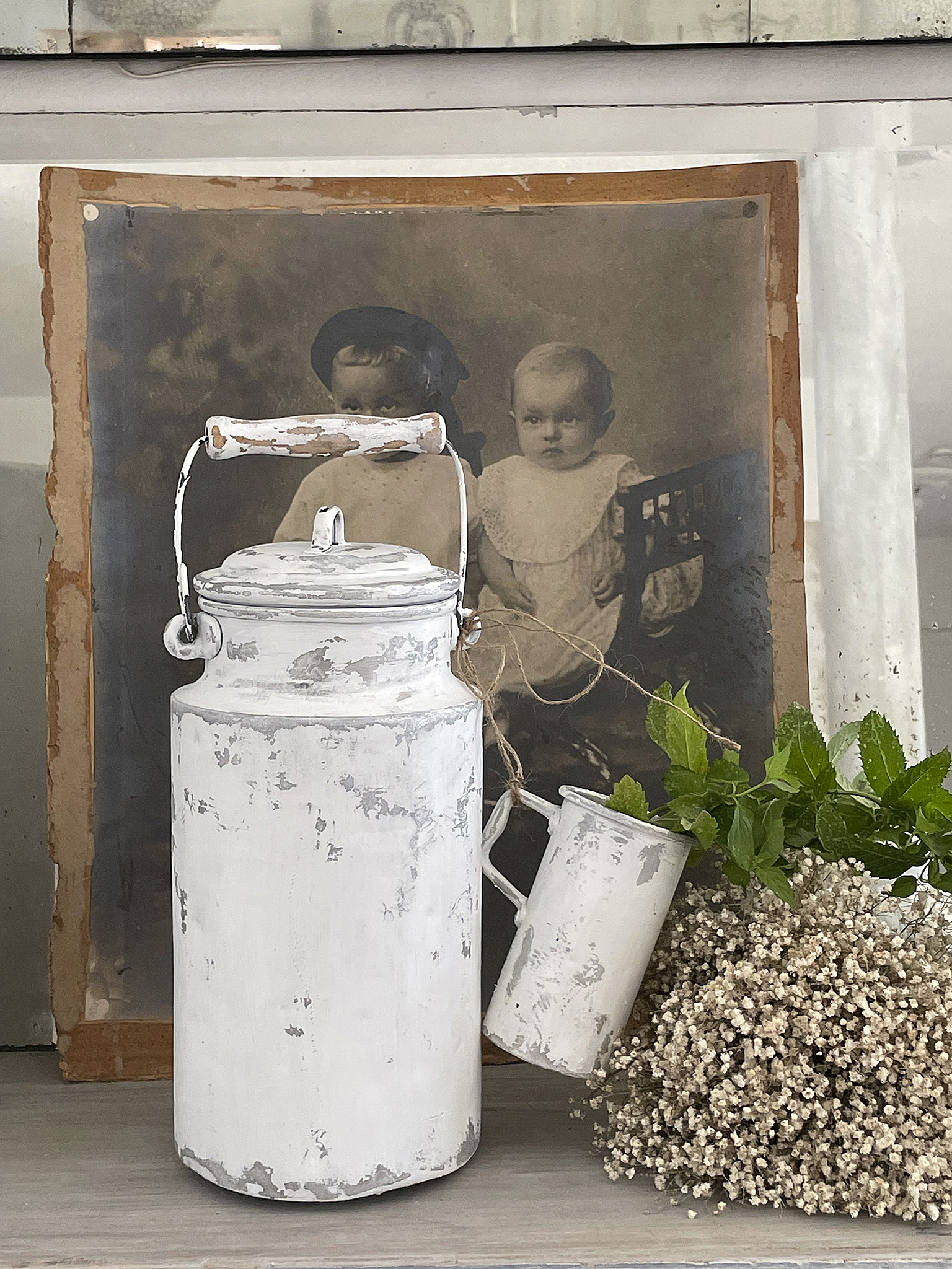 Vintage Milchkanne mit Dekoration und alter Fotografie im Hintergrund Weiße Vintage Milchkanne neben einer kleinen Tasse und frischen Kräutern, davor eine alte Fotografie von zwei Kindern.