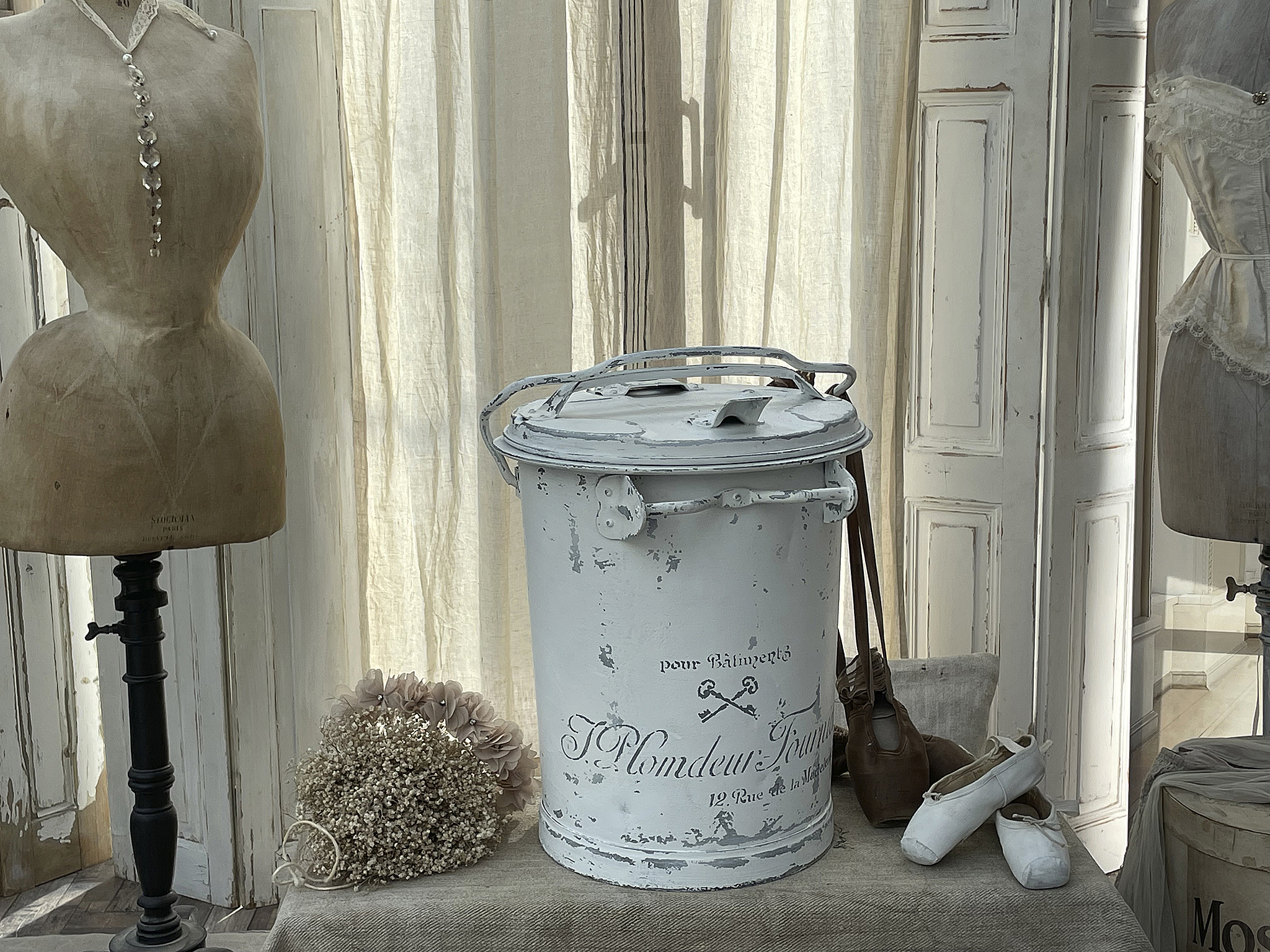 Weißer Vintage-Mülleimer mit dekorativen Accessoires Weißer Mülleimer auf einem Tisch, umgeben von einer Schneiderpuppe, Trockenblumen und Ballettschuhen.