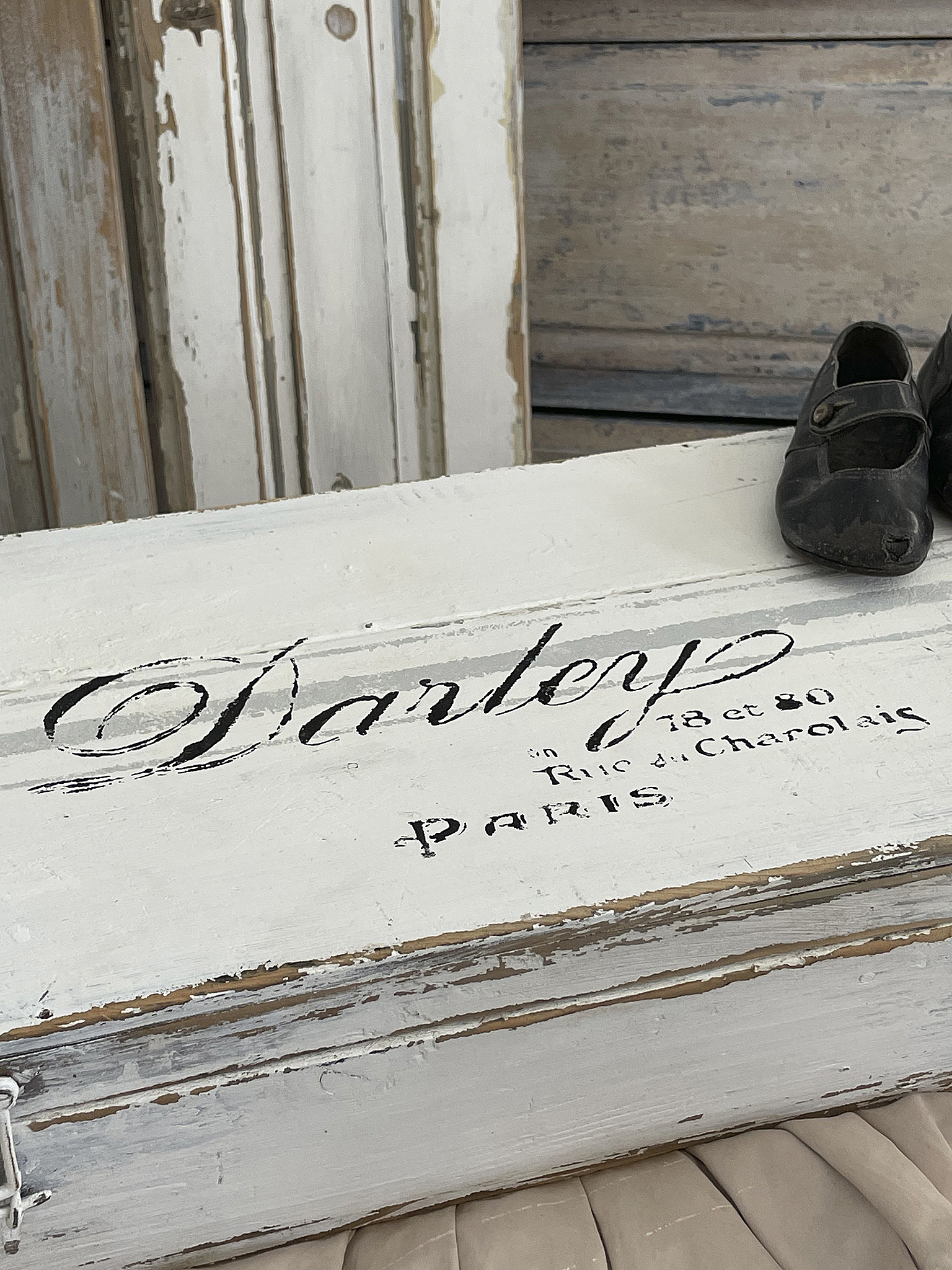 Vintage Holzkiste mit Schuhen und Paris Schriftzug Eine alte Holzkiste mit dem Schriftzug Danley und einem Paar schwarzer Schuhe, im Hintergrund eine beige Stoffoberfläche.