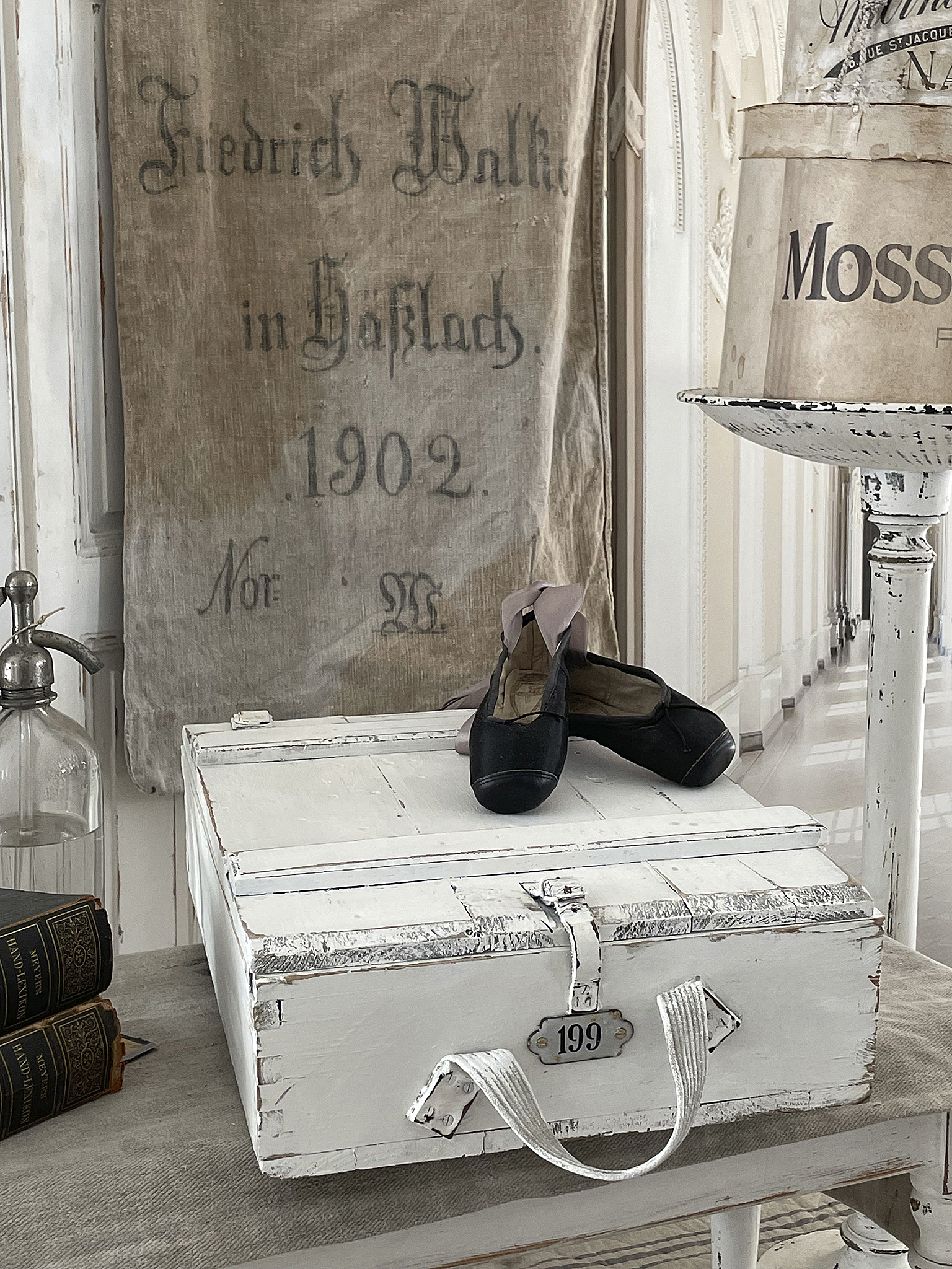 Vintage Aufbewahrungskiste mit schwarzen Schuhen und historischem Leinensack Weiße Holztruhe mit der Nummer 199, daneben schwarze Schuhe und ein alter Leinensack von Friedrich Walke aus 1902
