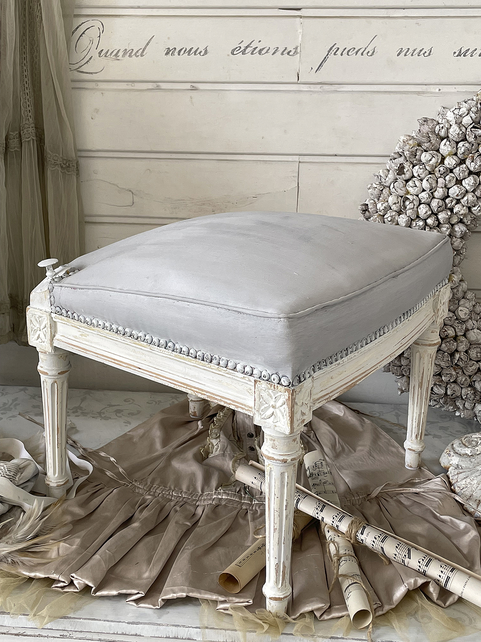 Eleganter Hocker mit grauem Polster und kunstvollen Details Eleganter Hocker mit grauem Polster, kunstvollen Beinen und Stoffen im Hintergrund