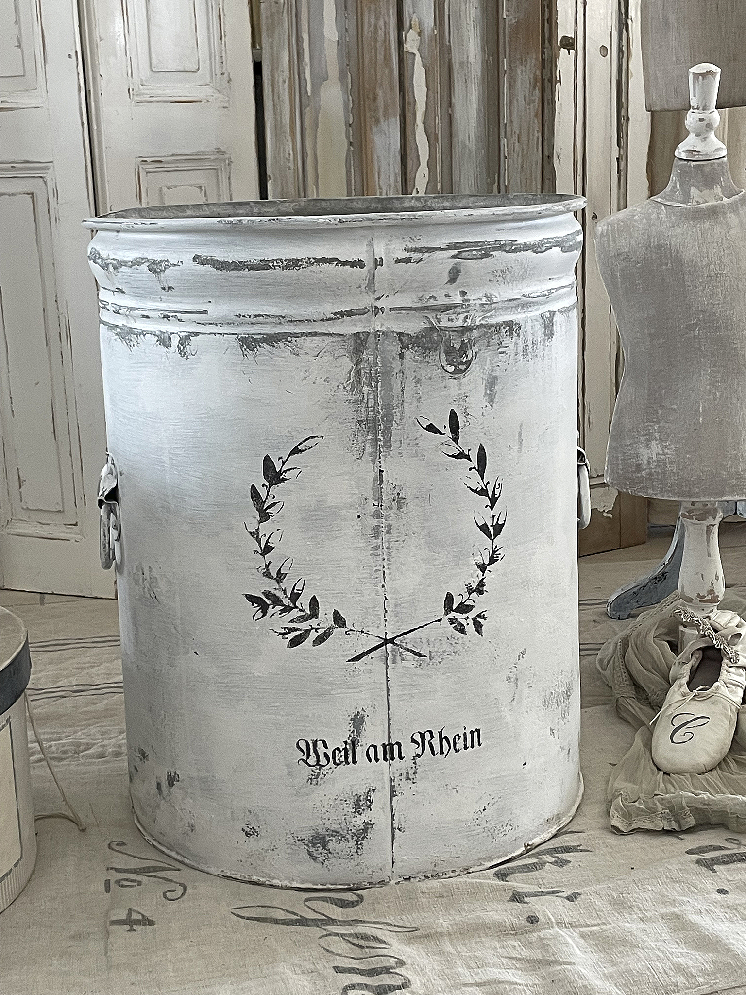 Dekorativer Metallbehälter mit Schriftzug „Weiß am Rhein“ und Pflanzenmotiv Metallbehälter in Weiß mit dekorativem Design, Schriftzug „Weiß am Rhein“ und Pflanzenillustration.