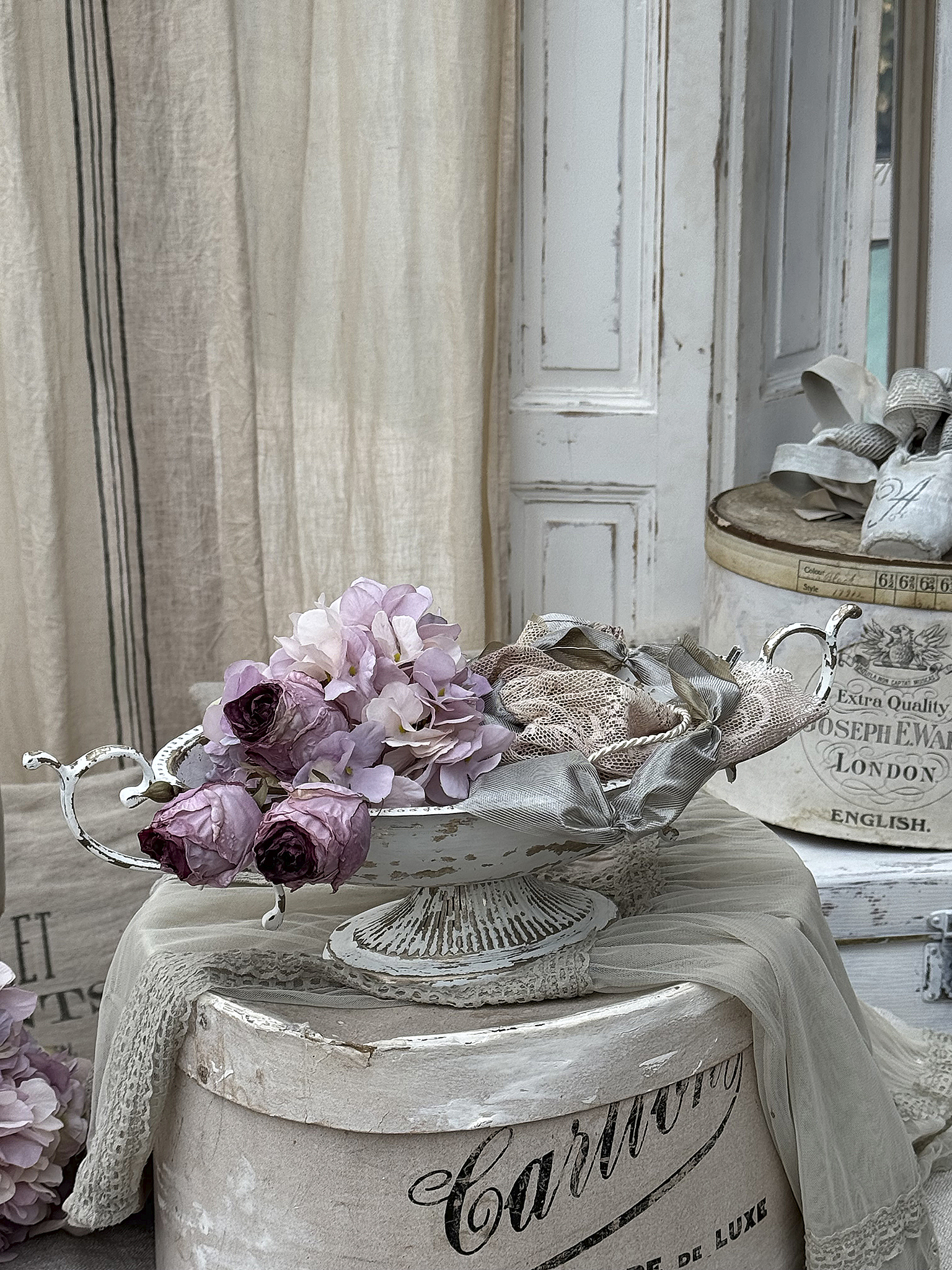 Schöne Schale mit rosa und lila Trockenblumen auf einem Tisch, umgeben von Textilien und alten Kisten.