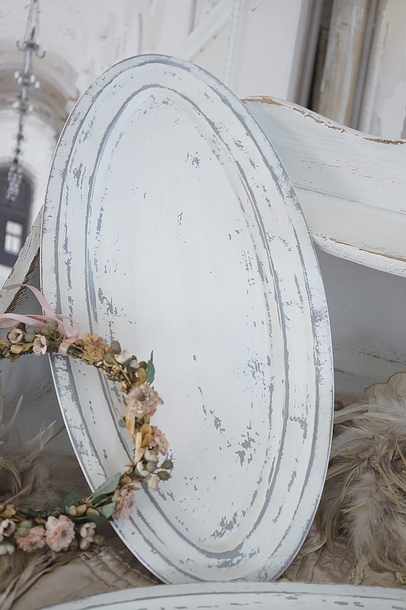 Weiße, ovale Servierplatte mit abgenutztem Design und einer Blumenkrone aus Trockenblumen.