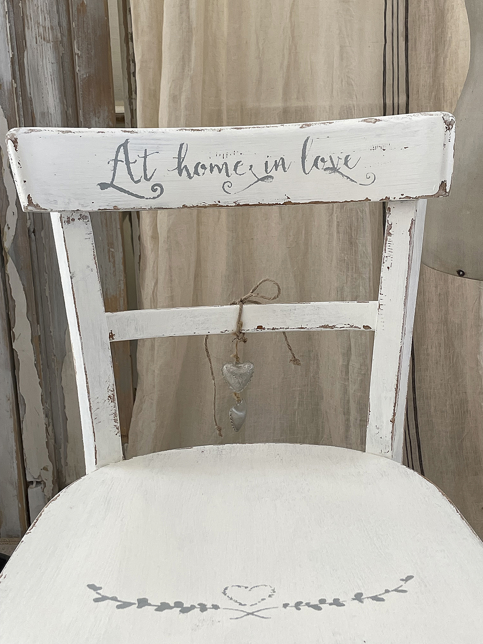 Vintage Holzstuhl mit handbeschrifteter Aufschrift und herzförmigen Anhängern Weißer Holzstuhl mit der Aufschrift 'At home in love' und dekorativen herzförmigen Anhängern