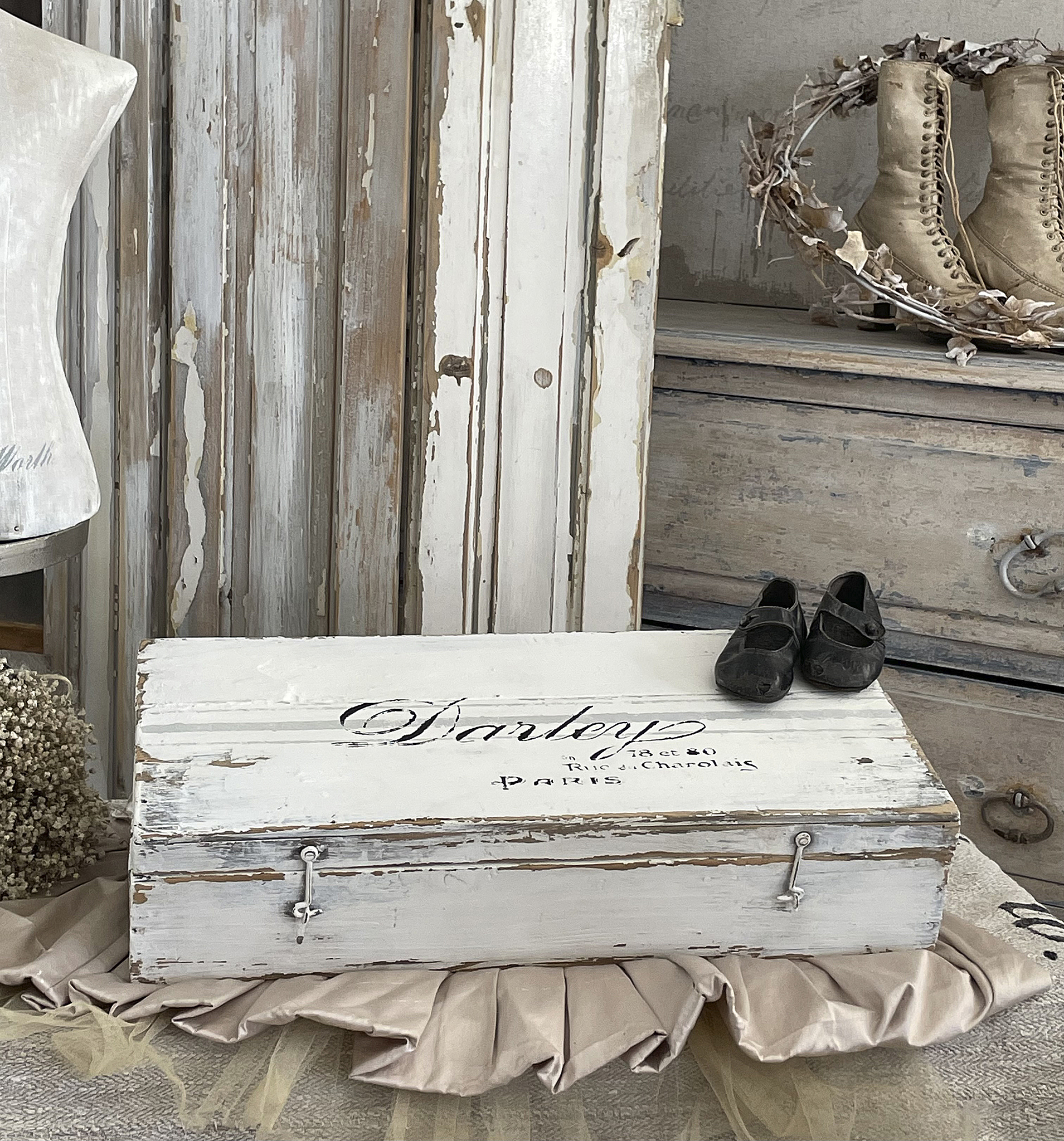 Vintage Aufbewahrungskiste mit Schuhen und dekorativem Hintergrund Weiße, vintage Aufbewahrungskiste mit dem Schriftzug 'Dartrey' und schwarzen Kinderschuhen auf einer eleganten Unterlage.
