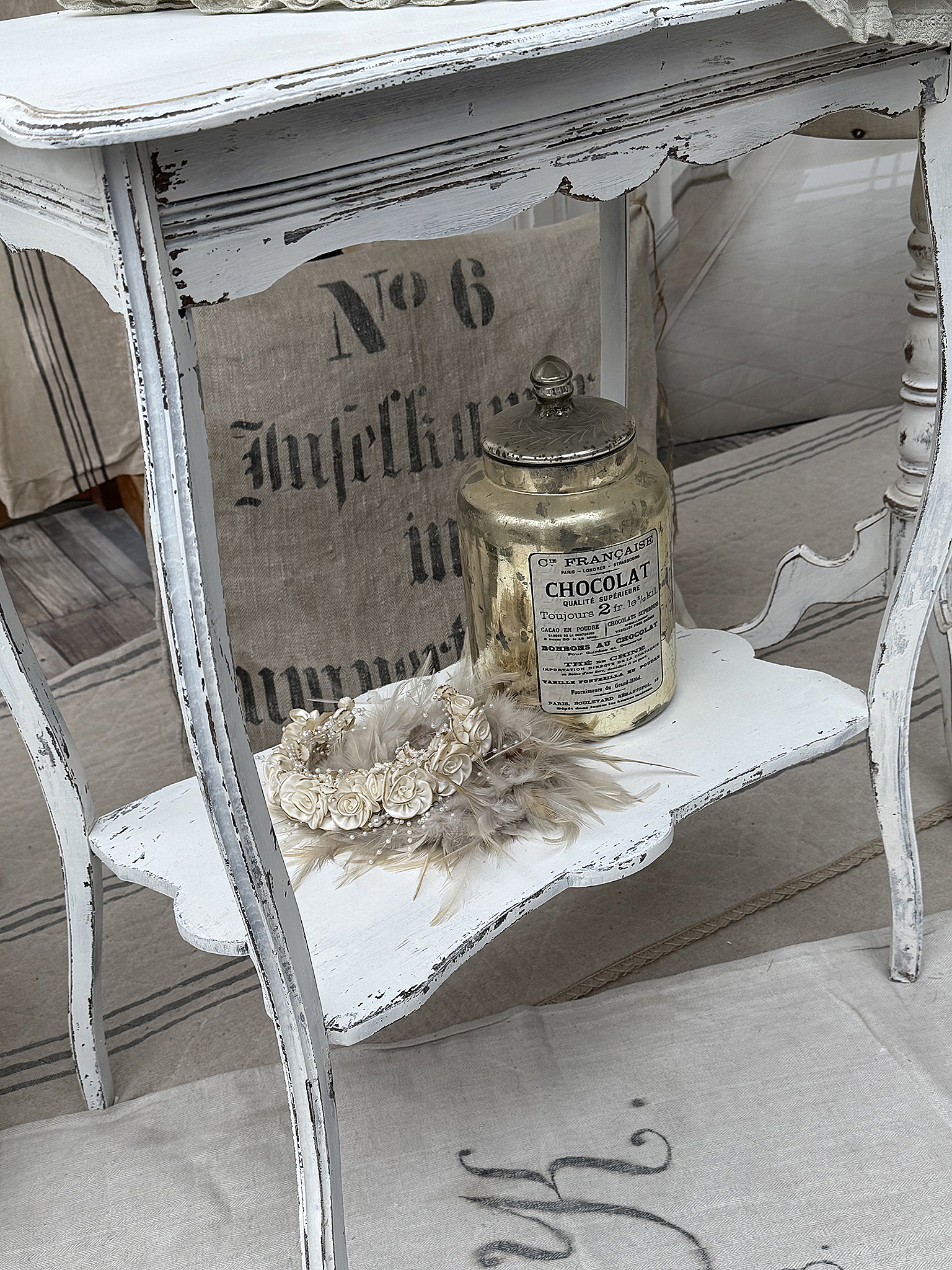 Vintage Beistelltisch mit goldener Schokoladendose und Blumenkranz Detailansicht eines weißen Beistelltisches mit goldener Schokoladendose und dekorativem Blumenkranz