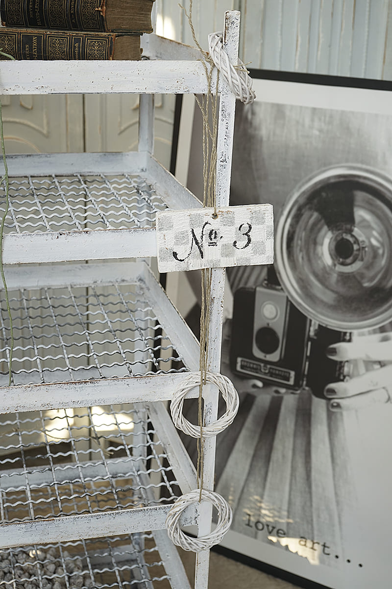 Detailaufnahme eines weißen Regals mit Vintage-Büchern, einem Schild mit der Aufschrift Nr. 3 und einem schwarzen Kunstbild im Hintergrund.