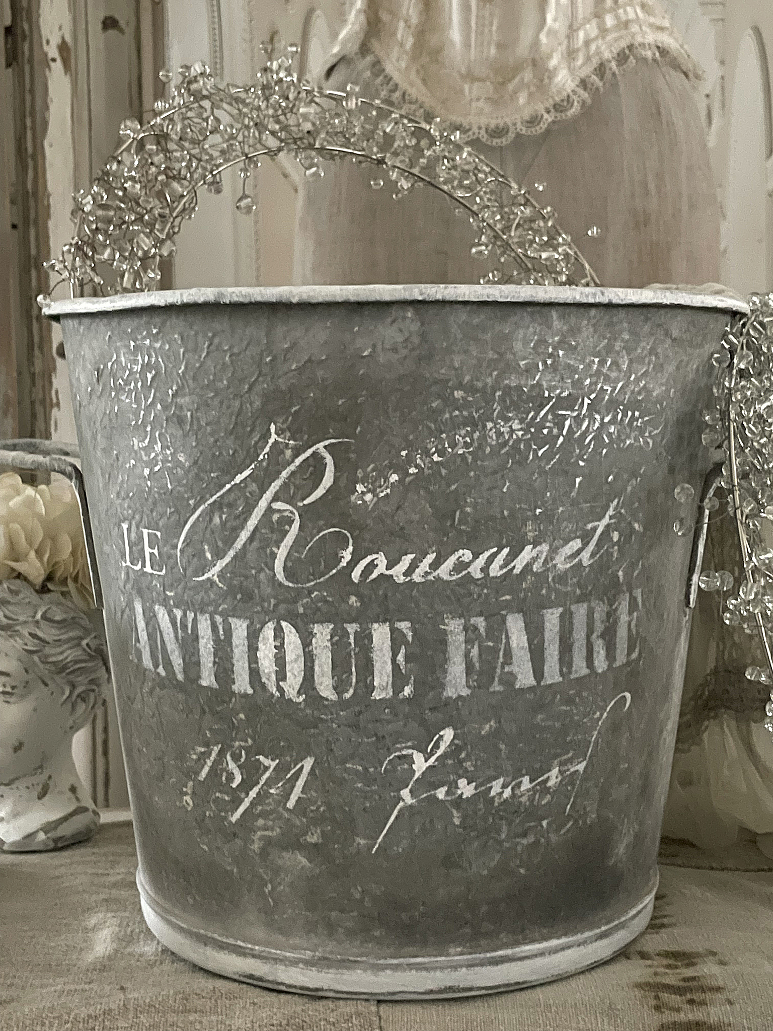 Antiker Metall-Eimer mit nostalgischer Aufschrift und Dekoration Antiker Eimer aus Metall mit Schriftzug 'LE Roucunet ANTIQUE FAIRE', geschmückt mit drahtiger Blüten-Dekoration.