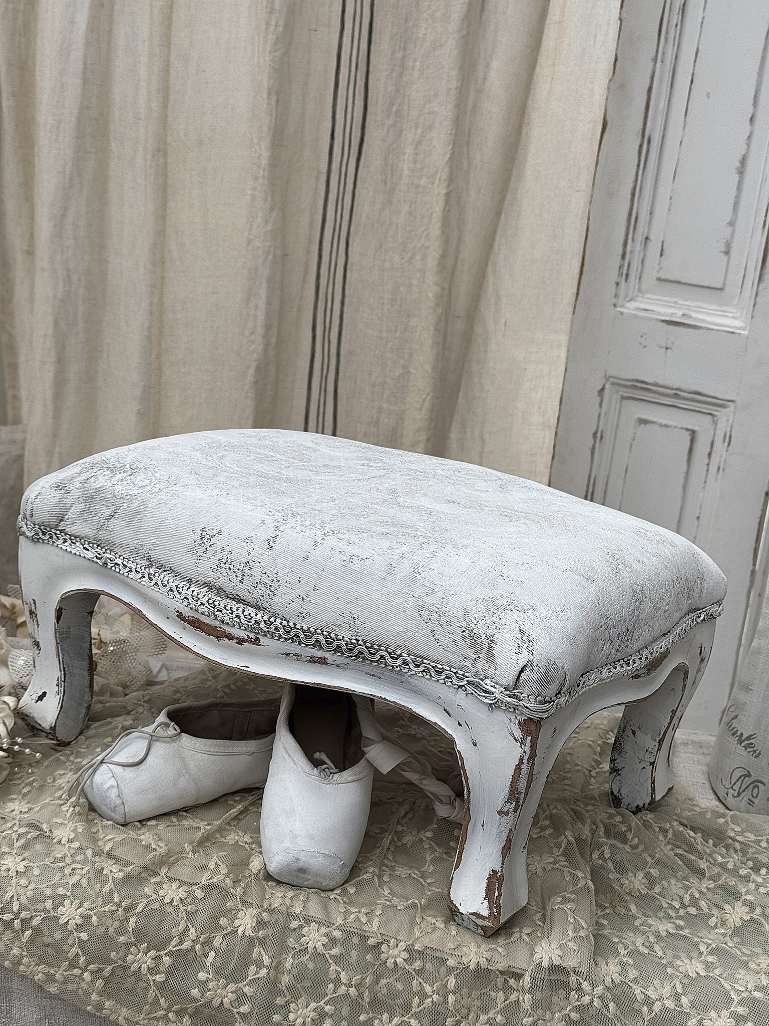 Eleganter, weißer Hocker mit Ballettschuhen, rustikaler Charme Weißer Hocker mit abgenutztem Bezug und Ballettschuhen darunter, ideal für ein stilvolles Zuhause.