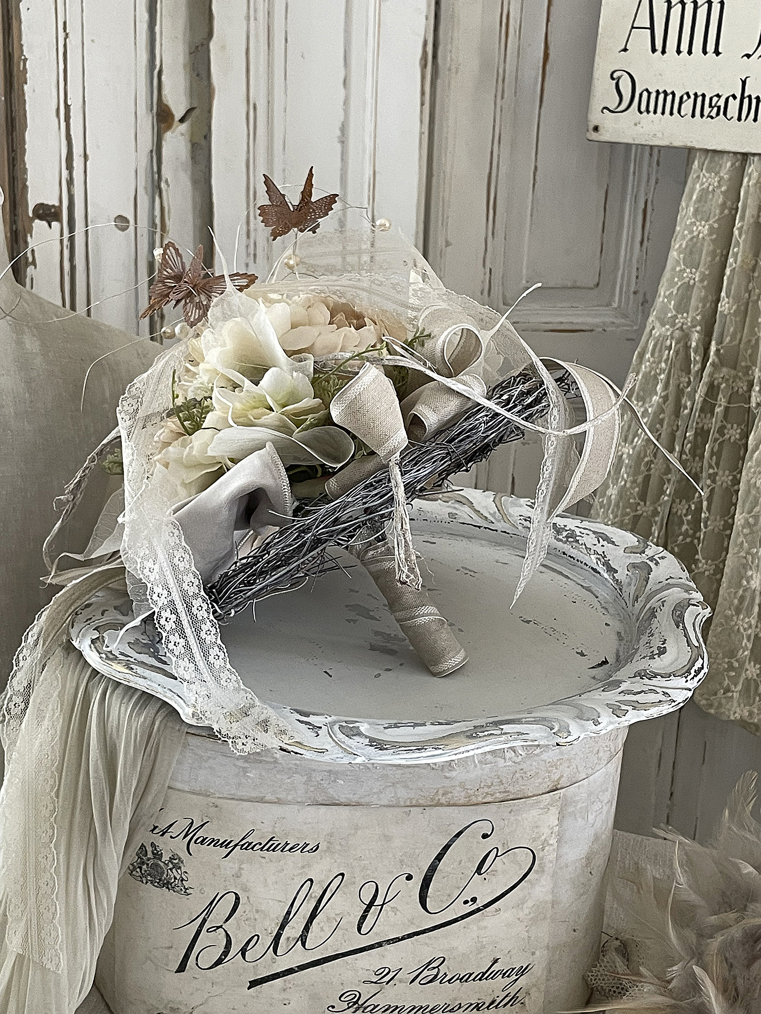 Eleganter Blumenschmuck mit Schmetterlingen in einem Vintage-Behälter Dekorativer Blumenschmuck mit beigen und weißen Blumen, verziert mit Schmetterlingen und Ribbons, auf einem antiken Behälter