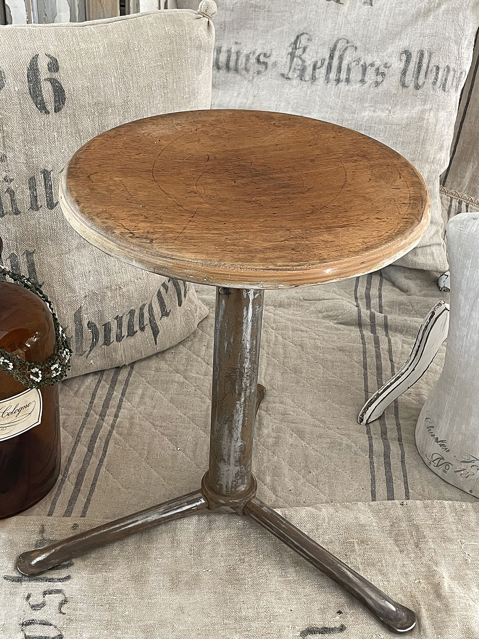 Kleiner runder Holztisch auf einem Metallgestell, umgeben von vintage Kissen und einer Flasche.